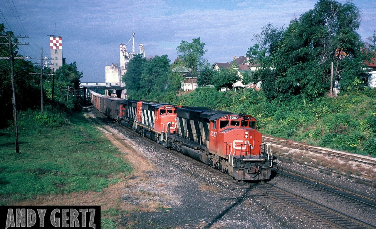 A CN eastbound led by CN SD40-2(W) 5357 is approaching the VIA Rail station in Woodstock, Ontario on the Dundas Subdivision. Trailing is GP40-2L(W) 9454 and an SD40. Also, the Purina Mill in the background has a cut of cars in one of their sidings.