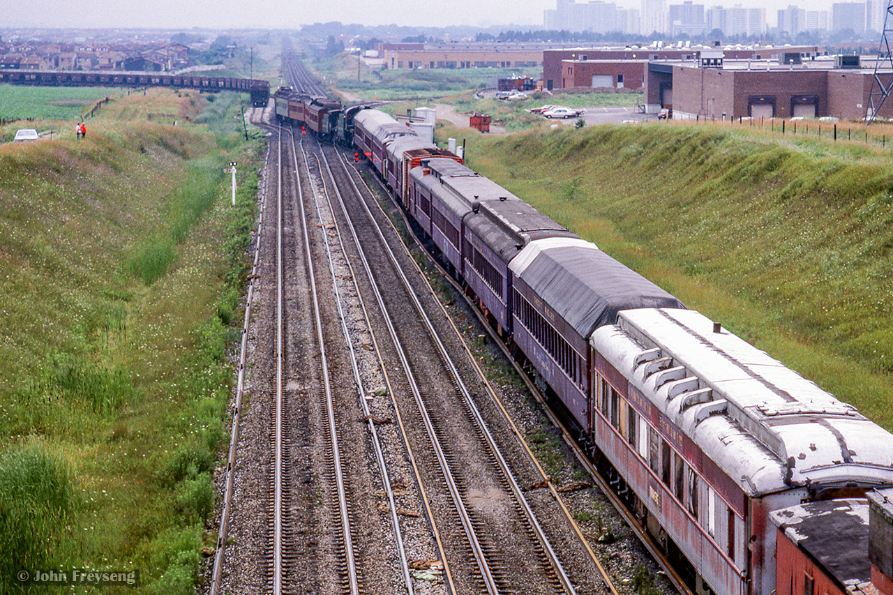 Negotiating the crossovers at Snider, a special equipment move for the Ontario Rail Association is underway from Toronto - Beeton via Allandale on Friday, August 1, 1986 for the new South Simcoe Railway. With CN engineer Gary Murray at the throttle of the pair of 4200-series GP9s, the move is well in hand for the trip. Following the power is four heavyweight passenger cars, ex-CPR 4-4-0 136, ex CPR 4-6-0 1057, three more heavyweight passenger cars, CLC DTC 22, a combine, a boxcar, another combine, three more heavyweight passenger cars, ex CPR wooden van 435398, a wooden MoW boxcar, ex CPR wooden van 436954, and in service CN van 79418.  Off to the left, note the empty hoppers on the Newmarket-York north connecting track.  These cars made up the stone train, operating between Nelson AggregatesUhtoff quarry/a> and Nelson's site on the Uxbridge Sub at Milliken.

Engineer Gary Murray has provided us with a first hand account of the move below:
Knowing this move to Beeton was in the works, I told the trainmaster that I would volunteer to take the train to Beeton on my own time. There was no precise date arranged yet but I kept waiting hoping they would use me. As time drew on I was afraid I would miss out because I had scheduled vacation quickly approaching. In the end I was given the assignment even though I was actually on vacation. I took a little slag over that from the other crews.

The original plan was to use two CNR F7A's as power so I was disappointed to see we were getting two GP9's. A Mechanical Supervisor was riding the 136 to closely watch for problems. We were speed restricted and stopped numerous times for walk around inspections. At Allandale we put the caboose on the north end of the train and ran around it. There was a huge crowd of people waiting for us. Old railroaders from the steam era and curious towns people. Many wanted to, and did, climb onto the equipment to rekindle old memories. After a good inspection and complete brake test, we left a crowd of admirers behind.

There were many more admirers enroute to Beeton as we stopped at many crossings to flag our way over. It had been a long time since any train had been over the old Milton Sub. Arriving at Beeton, once again we cut off and ran around the train so we could shove it over the crossing and leave it there south of the 8th line. There was a bit of an upgrade with poor rail conditions there causing our locomotive to struggle pushing this train. It was a characteristic of these locomotives to drop a part of their power when the locomotive was about to slip and then quickly reapply it. This was causing the slack to run out and then we would bump into the train. This happened a few times and I was concerned that we might upset some part of the consist. We eventually made it over the crossing and tied the train down. We then ran van hop back to Mac Yard . This was the last CNR move on this track.

As a footnote, our crew had a personal letter placed on our file commending us for our handling of this train. This sort of thing was so unusual that I considered having the letter mounted in a gold frame.

Scan and editing by Jacob Patterson.