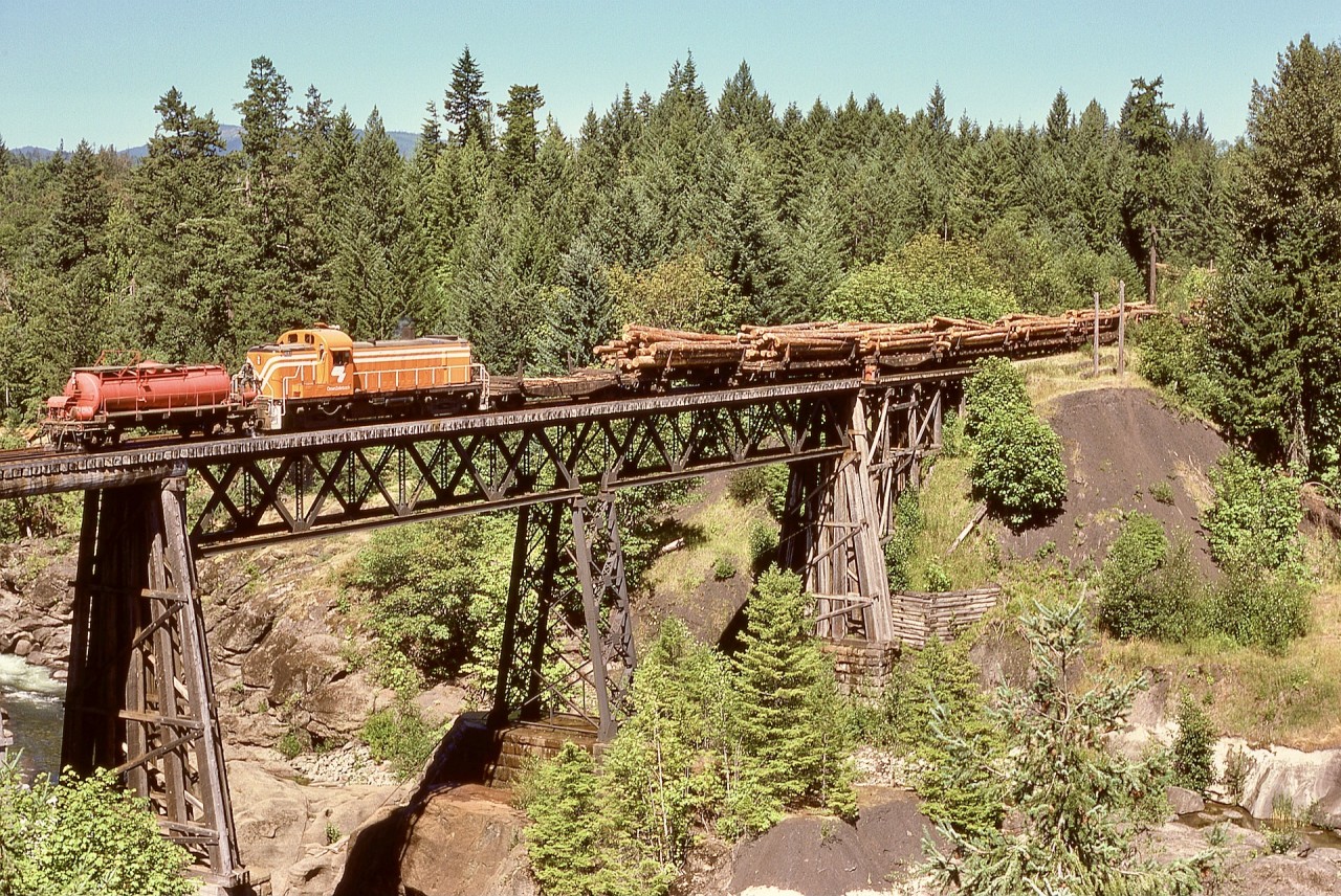 This is intended as a direct comparison with my post last week which showed CZ 8427 and log loads eastward in early morning sunshine on the same bridge seven years later.  This one, with ALCo RS-3 4097 with an active water car (note the brakeman has the flush valve open to discourage any brake shoe sparks on the wooden deck) was taken on the second trip of the day around 1100 PDT on Monday 1974-07-29 (just ten months since 4097 was painted orange with white stripes), with notably less pleasing lighting than the first trip, and was my first try at climbing the tall tree that became a favoured camera perch.  Within the limits of a 35mm camera and 45mm focal length lens, the river had to be omitted to include the tree line behind.

Often I wondered while waiting up that tree what would happen if something went wrong during my descent, as the crew would not have seen me, but would pass my truck (which they knew well) at the nearest road access half a mile further on.
