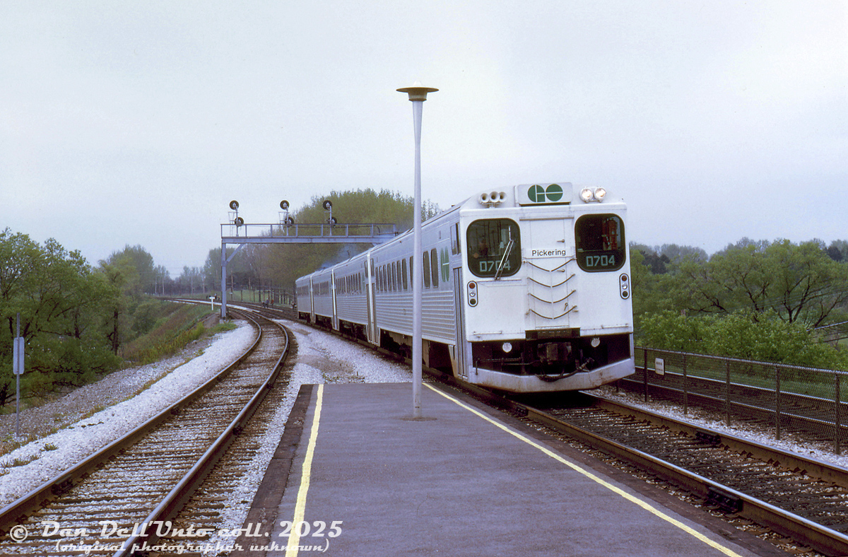 A 4-pack of GO Transit self-powered single level cars, lead by D704, arrive eastbound at Long Branch GO station in Toronto's west end. The consist is bound for Union Station downtown, and ultimately Pickering GO Station at the other end of the Lakeshore line. Some people linger on the tracks in groups past the bridge, possibly railfans on an unknown fantrip or outing trackside.

Along with all the single-level RTC-85 commuter cars GO purchased from Hawker Siddeley Canada for their startup in 1967 (and subsequent expansions), GO purchased 7 self-powered versions, dubbed RTC-85SP (and RTC-85SP for two units with dual cabs). They were intended to provide off-peak service on the Lakeshore line. Due to reliability issues, they were depowered and turned into cab cars in the mid-70's.

Original photographer unknown, Dan Dell'Unto collection slide.
