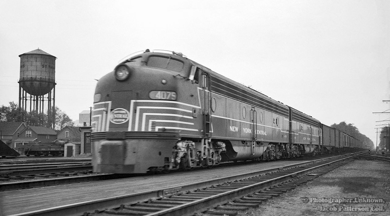 Behind a pair of lightning-striped EMD E8As, train 50, the eastbound Empire State Express, roll into St. Thomas at 10:25h if on time.  Lots of headend traffic makes up nearly half of this train.  At left, pieces of work equipment can be seen, including the tip of a wedge plow, and a small flatcar with parts atop it.  At right, the electrified connecting track of the London & Port Stanley Railway, running from the north side of the CASO station, west towards BX Tower, just visible in the distance, where it would swing north to connect with the L&PS mainline.

At left is the St. Thomas water tower, built in 1914, and holding the title of Canada's largest steel water tower, and second largest steel tank in North America.  It was demolished in 1993.   Photos taken from the tower in 1975 show various scenes of the town, including the railway.  Take a look at these pics here. 

Original Photographer Unknown, Jacob Patterson Collection Negative.