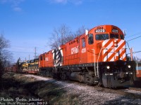 CP Rail C424's 4229 and 4219 head southbound on train #90 "The Moonlight", rolling through downtown Brampton in the afternoon on the way to Streetsville. They've just crossed Queen Street West and the former station site, and are approaching Jessie Street crossing near Mile 7.5. In tow at the head end are two flatcars of brand new Caterpillar machines, built at the Caterpillar Brampton assembly plant at Sandalwood Parkway and Hurontario Street, and lifted <a href=http://www.railpictures.ca/?attachment_id=44911><b>from their rail siding</b></a>. The first car appears to have three log skidders, the second a wheel loader and a log skidder.<br><br>Caterpillar's Brampton assembly plant opened in 1983 at the northwest corner of Hurontario and Sandalwood in the community of Heart Lake, and built small rubber-tired machines, including some of Cat's 900-series wheel loaders (e.g. the 926), IT-series Integrated Toolcarrier loaders (IT18/IT28), and 500-series logging skidders (518/528). It had a <a href=http://www.railpictures.ca/?attachment_id=56690http://www.railpictures.ca/?attachment_id=56690><b>rail spur and runaround</b></a> on the property to ship parts in and completed machines out. Cat had had another plant in Missisauga (Dixie, off the CP Galt Sub) that formerly did this manufacturing, but after assembly work was moved it continued to supply the new Brampton plant with parts for machine assembly. In the mid-80's, it was announced the Mississauga parts plant would be phased out and operations consolidated in Brampton.<br><br>By the late 80's, things weren't looking good for the Brampton plant. News sources at the time mention production at Brampton had declined 50% since 1988 partially due to the economic downturn, and more than 80% of the plant's production was being shipped to the US and also overseas (on the flip side, <a href=https://www.upi.com/Archives/1991/04/12/Union-objects-to-Caterpillar-closing-Canadian-plant/7078671428800/><b>CAW union leader Bob White countered</b></a> with Cat's overall sales outside the US made up 55% of the company's total sales at the time, despite Cat only having employment of 30% outside the US).<br><br>Cat made the decision to close Brampton and move production to a new factory in Raleigh, North Carolina, which according to officials had a lower cost of manufacturing. The Brampton plant, having undergone a significant modernization with new equipment prior to the decision, had that new equipment shipped to Raleigh (it may also be relevant to note the Brampton plant was unionized, and the Raleigh plant was not (right-to-work state)).<br><br>The Brampton plant was closed in early 1992, and the property sold to the Woolworth/Woolco department store chain for warehouse use (that eventually became Walmart Canada when they bought Woolworth Canada to enter the Canadian market in 1994).<br><br><i>Peter Jobe photo, Dan Dell'Unto collection slide.</i>