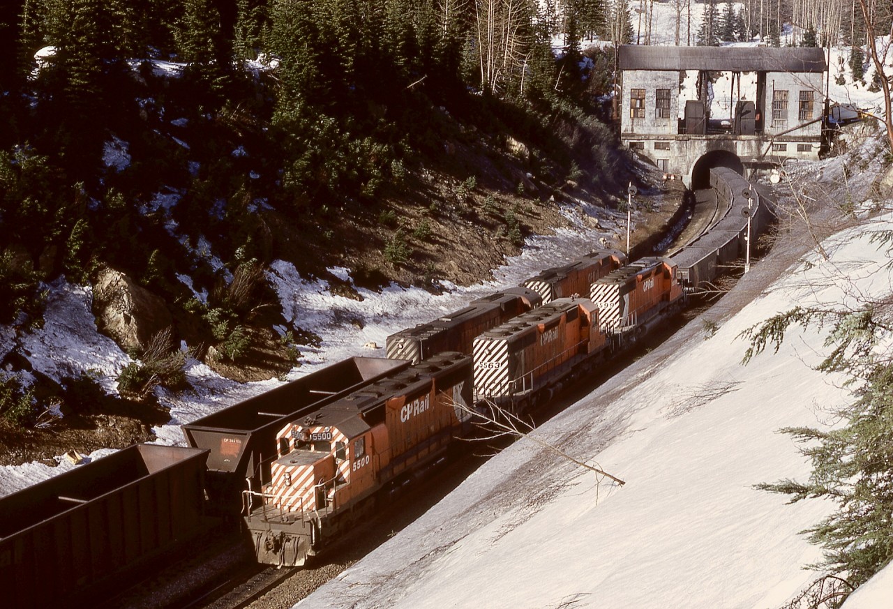 A motive power transition in progress is documented in this view at the west portal of the Connaught Tunnel, with two MLW M-630s, 4551 and 4557, on a train of empty rotary-dump coal cars heading east meeting two GMD SD40s, 5500 and 5563, plus SD40-2 5599 on a westward train of loaded coal cross-hopper cars (likely from Coleman, AB) on Tuesday 1974-04-23 at 1543 PST.