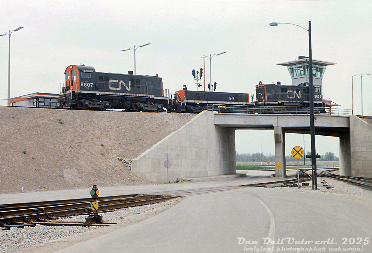 Clean ballast, fresh concrete, and newly converted hump power are all present in this view inside CN's Toronto Yard, their new state-of-the-art freight classification yard opened north of the city on acres and 1000 acres of former Vaughan Township farmland. CN S13's 8607, 8624 and slug B3 glide over the crest of the dual hump, past the dual hump office, hump signals and access roads. The original hump power, 8600-series MLW S13's weighed more than the standard 8500-series units, were equipped with newer GE 752 traction motors, and were equipped with hump controls and some other features including the ability to power chopped-down slugs that were converted from retired MLW S3 switchers.

Bounded by the former hamlets of Edgeley, Concord, Maple and Sherwood, Toronto Yard was part of CN's "Toronto Bypass" project that diverted freight traffic north of the city via the new Halton and York Sub connections, and out of congested Toronto, thereby freeing up track time for GO Transit to start up commuter operations and take over part of CN's Mimico Yard for their own facilities (Willowbrook Yard).

Renamed MacMillan Yard in 1975, it continues to serve an important part of CN's network today, albeit with newer technology and hump power. The S13-slug-S13 sets eventually gave way to GP38-2's and newer GMD-built slugs, and today SD40-2 variants and slugs.

Original photographer unknown, Dan Dell'Unto collection slide. 

More Toronto/MacMillan Yard photos:
C Yard tracks: http://www.railpictures.ca/?attachment_id=51354
Aerial view: http://www.railpictures.ca/?attachment_id=33864
South end: http://www.railpictures.ca/?attachment_id=52917
Departure/Arrival leads: http://www.railpictures.ca/?attachment_id=52621
Diesel Shop (outside): http://www.railpictures.ca/?attachment_id=1670
Diesel Shop (inside): http://www.railpictures.ca/?attachment_id=20758