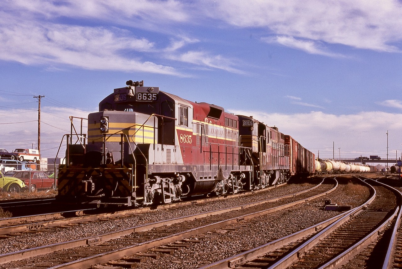 When the Fairbanks-Morse units on Canadian Pacific were approaching their end of service, one assignment remained strongly served by a few H24-66 Trainmasters — they were ideal for power, traction, and control for switching the hump yard at Alyth in Calgary, particularly as trailing units for modified GP9s.  Here, at the west end and approaching the 12th Street East interlocking tower (with the Blackfoot Trail overpass in the distance), CP 8635 and 8905 are slowly shoving a long string east over the hump on Wednesday 1974-10-02.

Ten years later, CP 8635, by then further upgraded and modified for non-hump duties and renumbered 1580, became one of the first assigned GP9 yard units at the Coquitlam Locomotive Facility in Port Coquitlam and part of my work world as an electrician.  It was among the last GP9s to survive in yard service there, through to the end of 2013.  Note its short hood is NOT a chopped GP9 hood, which would have a central vertical flat area on the nose, but is more like a GP35 short nose.  Two other Alyth hump units, 8633 and 8634 (which became 1579 and 1576) were similarly equipped.