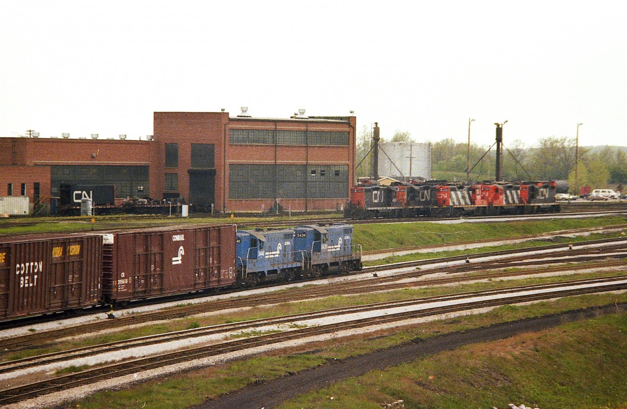 Conrail 5439 and 5441 are seen bringing over the daily transfer to Fort Erie. In behind is the once-busy CN shop complex. Lot of engines there, as usual, and at least five Geeps can be seen. Note on the left a shell of an unknown unit on a flat car. Some fairly heavy work was done in the shop as well.