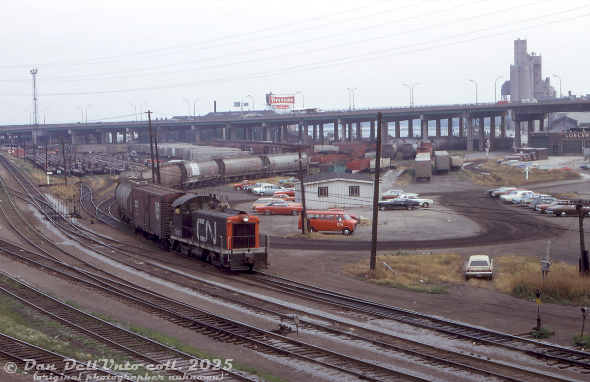 Downtown Toronto wasn't all passenger power and coachyards: CN SW9 7001 switches covered hoppers just east of Bathurst Street bridge (Cabin D interlocking), in a view looking south to the Gardiner Expressway and harbourfront area (today, this is all condos). 

The white building is CN's Yard office, the train is on the lead that curved south under the Gardiner and lead to the Central Harbour District and Queen's Quay harbourfront trackage, including Loblaws (part of their warehouse on the right) and the Canada Malting grain elevator (further back). Piggyback flats, boxcars and cylindrical covered hoppers pack the team tracks, storage tracks, piggyback ramps, and Cottrell Forwarding freight LCL platforms scattered here.

The lead on the right curving past Loblaws (note crossbucks) connects to the CP ex-TG&B Wharf Leaf that ducked out of view under Bathurst Street, passed through CP's Fez City Yard, under the Gardiner past Molson's, across the Oakville Sub at Cabin E interlocking, and on to the Liberty Village and Parkdale Yard areas (this was CP's western access to the downtown harbourfront industrial trackage).

On the left, tracks lead to Union Station, CN Spadina Roundhouse/Coachyard, and the "high line" mainline freight bypass.

Original photographer unknown, Dan Dell'Unto collection slide.