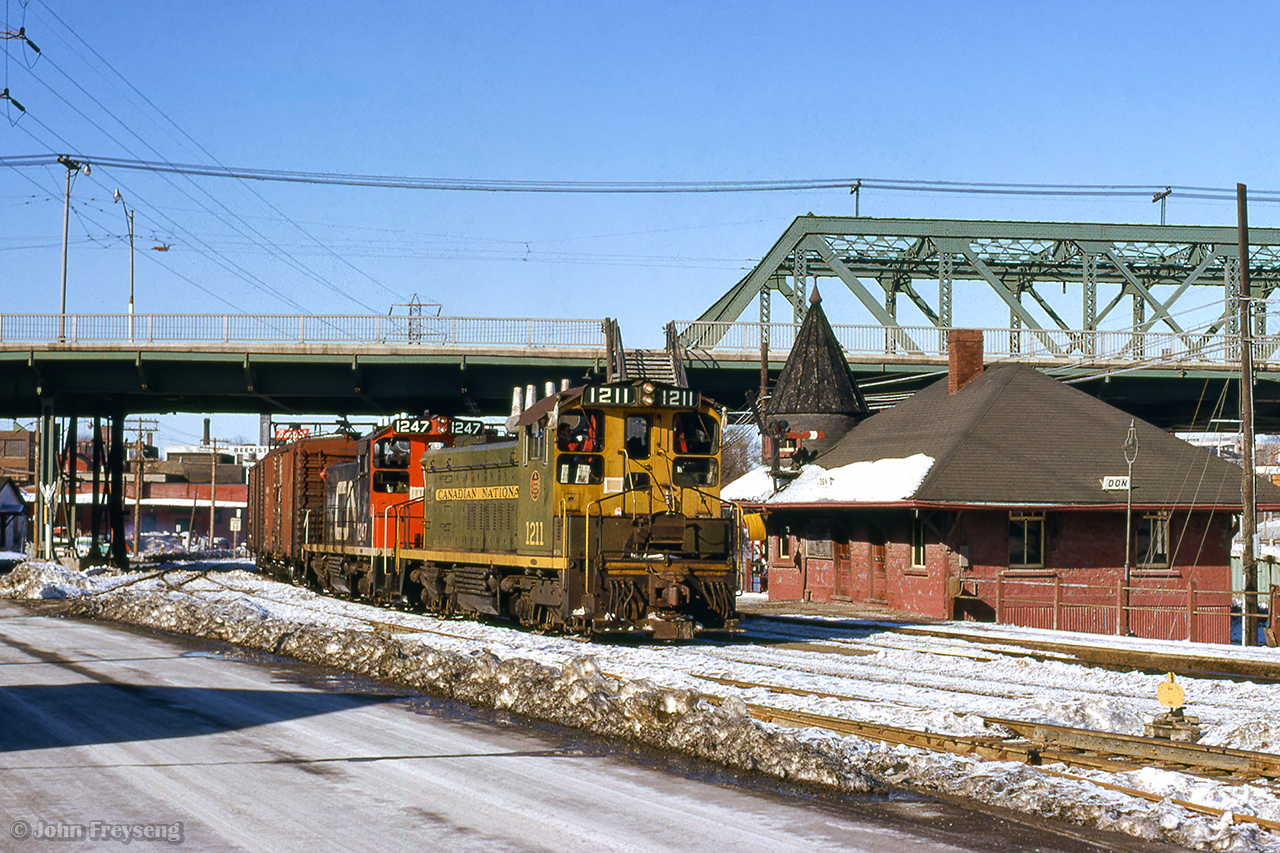 After meeting a Mimico - Toronto yard transfer <a href=about a mile north, the Toronto - Mimico yard transfer passes Don station beneath Queen Street.  Note the yellow flag hung on the station wall.  As this train order semaphore was only equipped with green and red lenses, a yellow aspect had to be created to allow trains to grab orders on the fly.  Red signal with yellow flag by day, or lantern on the platform by night, permitted this operation.

Scan and editing by Jacob Patterson.