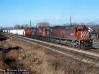 Filthy Big M's and fresh, clean tank cars: that's what sums up this acid train departing Kinnear Yard in Hamilton, caught by Reg Button passing the brick factory along Lawrence Avenue around Ottawa Street a few days before Christmas in 1988.<br><br>The end was near for CP's fleet of 6-axle MLW power, and M636 4714, M630 4554, and M636 4727 were certainly showing it: it's almost as if the cleaners at Agincourt Yard sprayed off the front of 4714, but then took a look at the rest of the unit and gave up on sight. And faded and sooty "CPR/ail" 4554 looks like she's seen a bit too much sun and a bit too much Alco smoke in trailing status. 4727 is the most presentable of the bunch, but its fuel tank streaked in leaking engine oil won't impress the shop crews much when they need to roll out another 55 gallon drum to keep her going.<br><br>All three were retired by 1993 when the final Big M units were shut down at St. Luc, in an official ceremony on December 23rd 1993. However, some that still ran (or managed to stumble off the shop track at St. Luc on their own) were unretired in 1994 due to a severe power shortage to run out a few extra miles, and by decree from higher up, were only run until major failures. None of the three pictured were so lucky, and some of the undermaintained, ailing units reactivated barely lasted a few months before being unceremoniously shoved back into the deadline. The lucky few managed to squeak their way into early 1995 before CP put the last ones out to pasture for good (except for the repowered M636-CAT 4711, that ran until 1998, and then was sold to Minnesota Commercial. A "Cat with 9 lives" maybe, as the Sartigan Railway has now acquired it).<br><br><i>Reg Button photo, Dan Dell'Unto collection slide.</i>