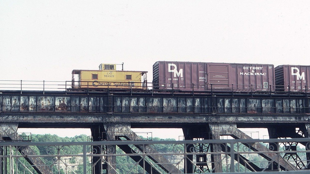 This photo of the C&O Chessie caboose was taken from the lower deck (vehicle traffic) of the Whirlpool Bridge over the Niagara River Gorge on US Independence Day, July 4, 1983, as we drove west into Canada. The Chessie train from Buffalo has left NY State and is entering Canada, headed westbound into Niagara Falls across the adjacent, upstream, Michigan Central Bridge.  The MC bridge is of course no longer used and the associated mainline tracks through Niagara Falls are no more. Those Detroit & Mackinac box cars were often seen.