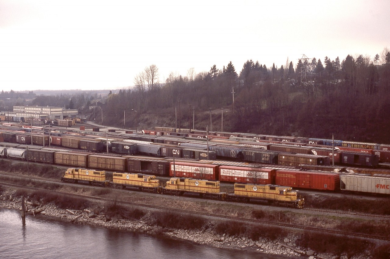 Since BC Hydro had a general restriction for only three of their four SD38 units on the headend of a train, catching all four working together was fairly rare.  For the Valley Freight from New Westminster to Huntington on Tuesday 1982-03-23, for reason(s) unknown to me, all four, BCH 382 + 383 + 381 + 384 (listed from right to left) were lined up, and as seen from the Queensborough road bridge over railway and Fraser River, are heading from the west end shoptracks (which are in Burnaby) at Trapp Yard to tie onto their train and head eastward.

BCH 381 is an SD38AC, and the others are SD38-2s, and all are EMD built.

The track between those units and the river is CN’s Lulu Island branch.