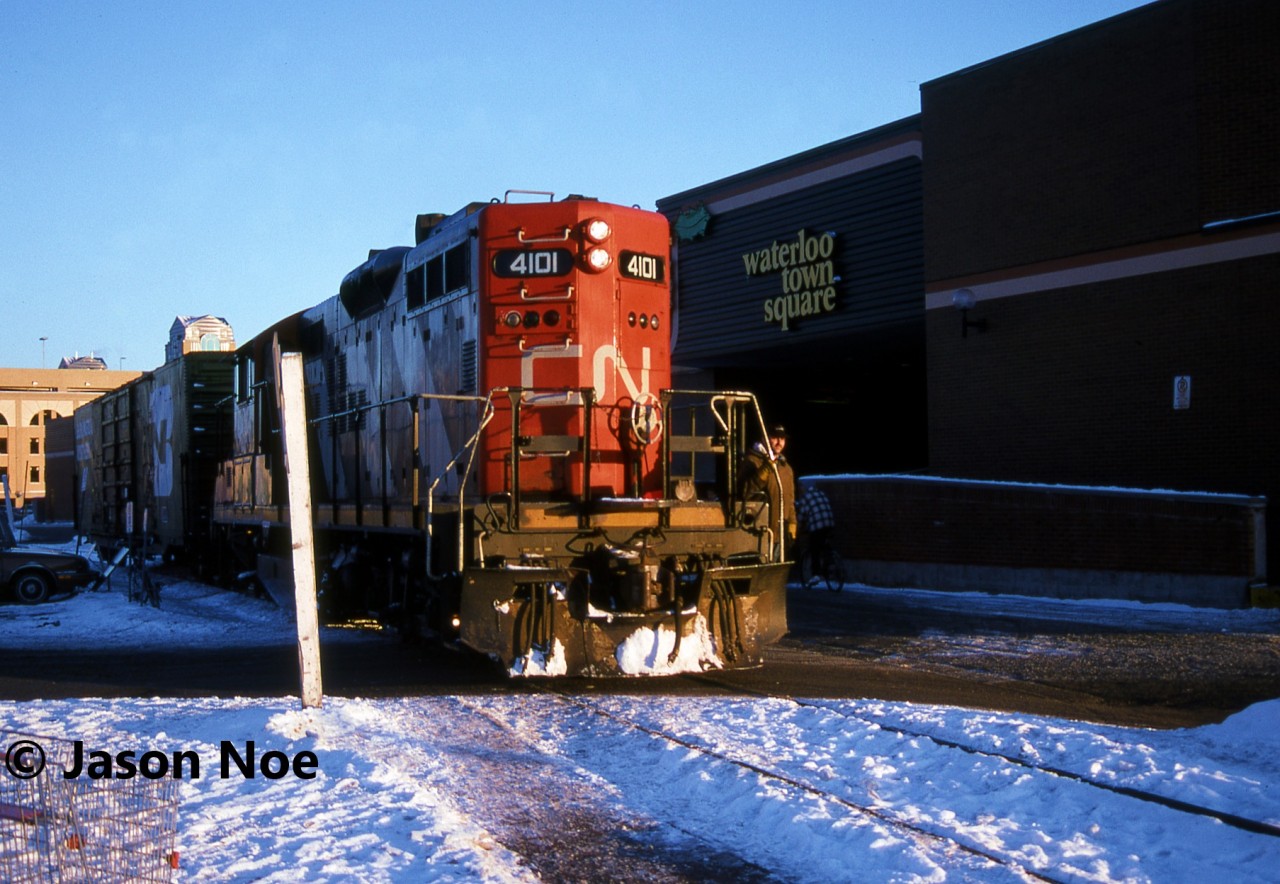 By the beginning of 1996, both CN and CP had completed the deliveries of their latest motive power. CN’s order of SD70I’s (5600-5625) and CP’s order of AC4400CW’s (9500-9582) had assisted both railways with easing some of their previous motive power constraints. While summer prior, CP had officially retired their last big MLW’s, they still had an active roster of RS18u’s and C-424’s operating in Ontario and Quebec. As well, despite the addition of the 83 new AC4400CW’s, the railway continued to lease hundreds of units through various companies, mainly Helm Leasing. CN continued to roster a small number of operating M-636’s, plus had their HR-616’s, M-420(W)’s and HR-412(W)’s working mainly in Ontario to eastern Canada. However, the company’s next big order was due to start arriving that summer, in the form of 105 new SD75I’s from GMDD in London, starting at CN 5626. Additionally, the start of the Canadian shortline takeover of CN and CP’s unwanted secondary lines and branchlines was still over a year away. Although, many had already fallen or were in the process of being abandoned. Two local examples were the CP Owen Subdivision from Orangeville to Owen Sound and the CN Newton Subdivision from Stratford to Palmerston, as well as the remainder of the CN Owen Sound Subdivision to Harriston.

Here, the Goderich-Exeter Railway (GEXR) takeover of the CN Guelph Subdivision was just under two years away as CN GP9RM 4101 leads one boxcar through downtown Waterloo on the Waterloo Spur. The 15:30 Kitchener Job was taking the car to Commonwealth Plywood in north Waterloo and would then return light power back to the Kitchener Yard.