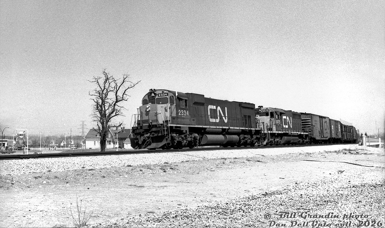 On a sunny October day in 1972, CN M636 2334 and SD40 5075 lead an extra freight off the Halton Sub at Burlington, about to enter the Oakville Sub and head west. The first few cars are passing over the Brant Street underpass.This photo was taken not too long after the Brant Street grade separation project (that took place in the late 1960's). Looking at 1965 and present-day aerial imagery it's hard to note the exact changes, but it appears Brant Street was realigned to the east, and the (closer) houses in the background were once along the south side of Plains Road East where Burlington Hyundai is today. The Sunoco may be the same one on the SW corner that was a Petro Canada until recently. Some more gas pumps appear on the NE corner, and beyond that housing along Leighland Rd (visible before the current plaza was built).Bill Grandin photo, Dan Dell'Unto collection negative.