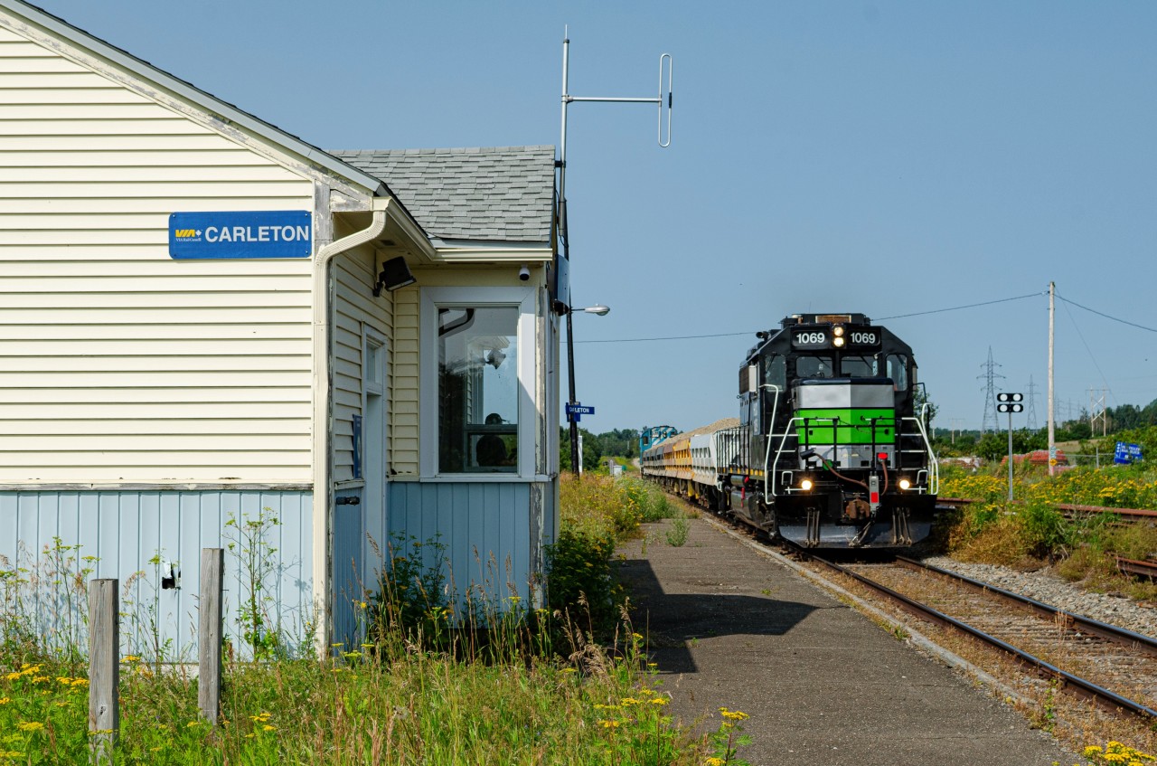 Leased GP38-2 1069 pulls a loaded ''Beluga'' train through disused Carleton VIA Rail station. These trains ran back and forth between Nouvelle and Maria in the summer of 2025, carrying crushed stones for the beach replenishment project in Maria. This project was handled by Construction Beluga, hence the train's nickname. On the tail end, leading the empty run that day was SFG 1849, the Amiral RS18u.
