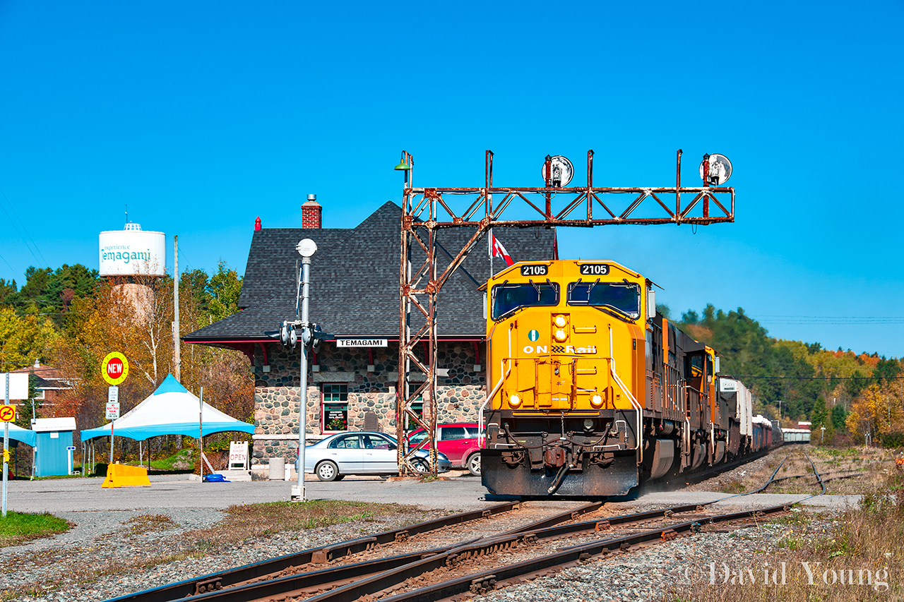 A large southbound freight rumbles past the historic station and under the old ABS cantilver signal bridge in downtown Temagami. Once a staple landmark, the old signal bridge finally came down in 2025 and the crossing got a face lift as well, outfitted with gates.