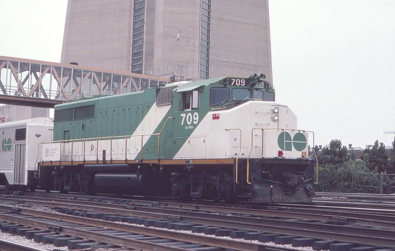 GO Transit 709 is seen with a commuter train west of Toronto Union Station in July of 1982. The concrete base of the giant CN Tower looms behind the GP40-2L