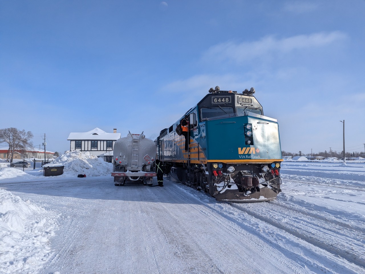 VIA Rail’s transcontinental train #1 pauses at Sioux Lookout under a crystal-clear winter sky as locomotives 6448 and 6457 take fuel. The snow is dry, white, and squeaks underfoot with every step — a sure sign of deep cold. Though the mid-afternoon light makes the scene look almost mild, temperatures sat at –28°C with a –38°C wind chill, making even a moment without gloves a test of endurance. Passengers step off briefly while the Sioux Lookout station anchors the scene at left.