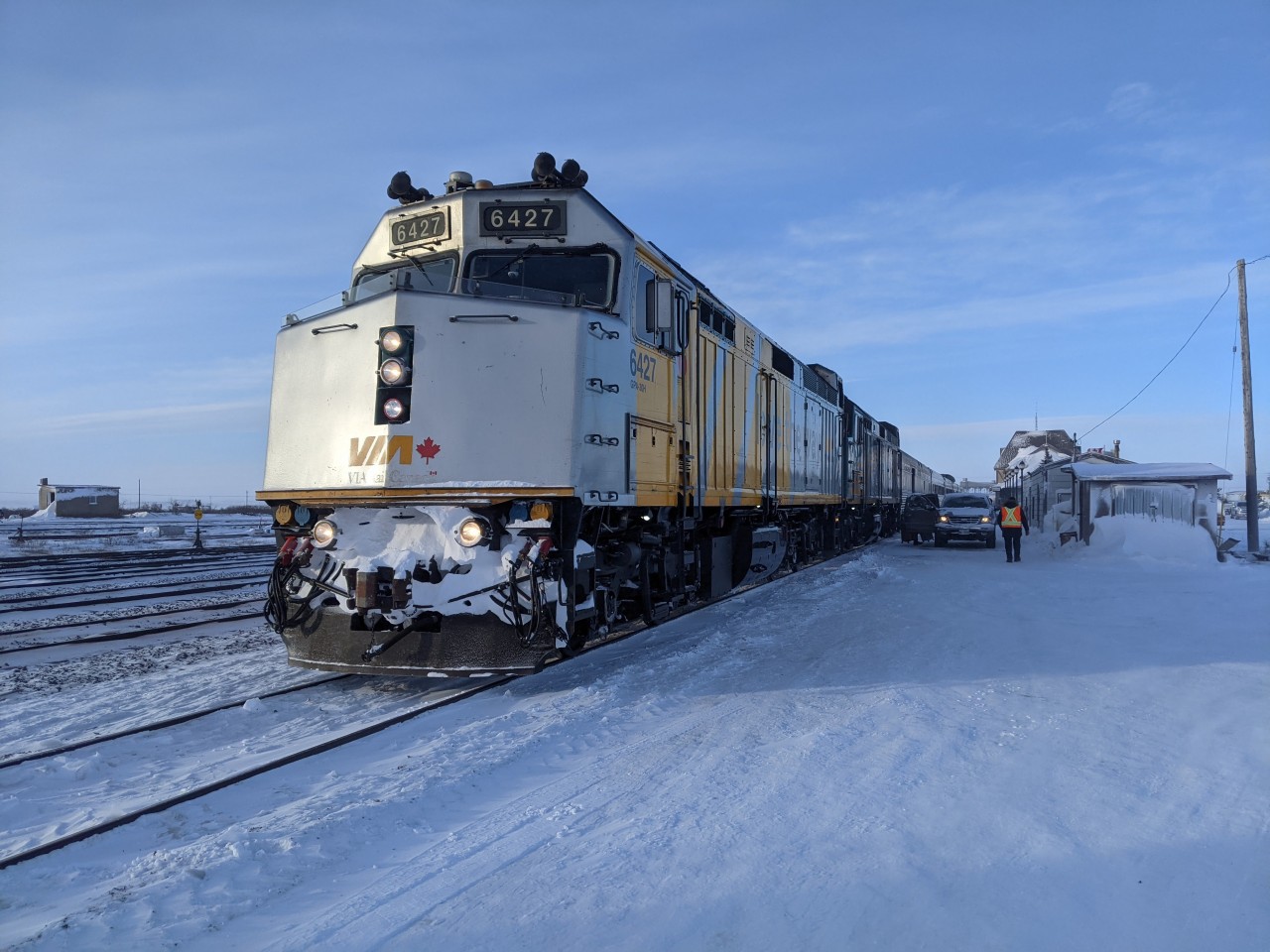 VIA Rail units 6427 and 6438 stand quietly in what little mid-day sunlight Churchill, Manitoba offers in late January. At just 1:00 pm the sun already hangs low in the sky, and the locomotives soak up every bit of warmth they can as temperatures sit at a brutal –28 °C, with a wind chill near –43 °C thanks to the wind off Hudson Bay. Train #693’s 47-hour journey north from Winnipeg carried it straight through a polar vortex, bringing exceptional cold and frozen sections of the train’s plumbing along the way. To the right, the roofline of the historic Churchill station (built in 1929) rises from the snow; to the left, multiple tracks lie silent, as empty as the platform itself. Passengers have long since retreated indoors, leaving only the photographer and a few hurried crew members finishing their duties at the end of the line before heading on rest — with a southbound departure back to Winnipeg just 12 hours away.