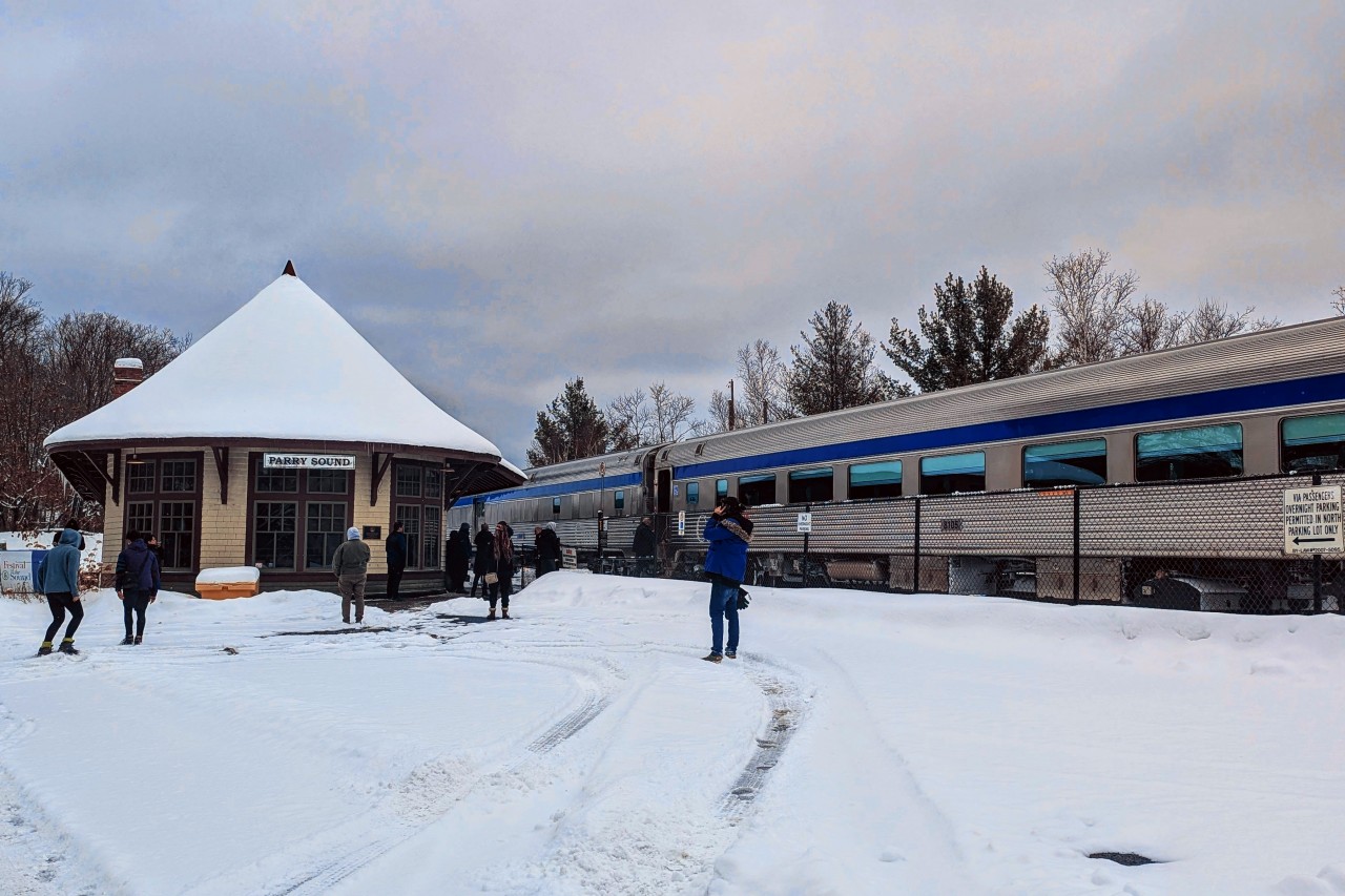 January 2026 at Parry Sound, Ontario. A portion of VIA Rail’s Canadian (Train 1, Toronto–Vancouver) stands during a scheduled fresh-air break, with the locomotives (VIA 6448 and 6457) just out of frame to the left. From this angle, the baggage car (8606) and first economy coach (8105) are visible as passengers detrain for their first opportunity to stretch their legs roughly five hours into the journey, having departed Toronto at 10:00 a.m. With no onboard Wi-Fi and limited cellular coverage along much of the route, the stop offers a brief chance to check phones, grab some fresh air, or — in the photographer’s case — dash out to document the train itself.

Photographic options here are limited by nearby residences, fencing, and the steep terrain, but the moment is anchored by the historic Canadian Pacific Railway station in the background. Built in 1907, the castle-like CP station ceased regular passenger service in 1990 and today houses the Festival of the Sound offices. VIA Rail continues to use this location, known operationally as Parry Sound South, for westbound service, while eastbound trains call at the former CN station elsewhere in town — a reminder of Parry Sound’s layered railway history.