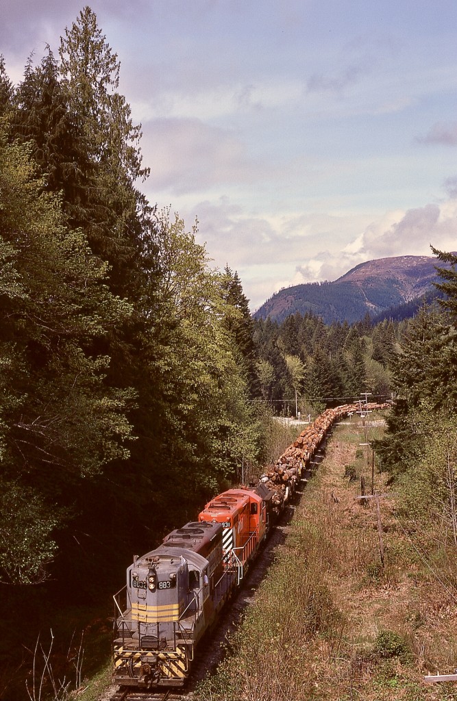 Canadian Pacific’s E&N Division on Vancouver Island handled trains of logs from Crown Zellerbach’s Nitinat Logging Division (hence the name Nitinat for those trains), eastward for 18 miles from Lake Cowichan to Hayward then 17 miles northward to the CZ log dump at Ladysmith. As seen from the last water tower at mileage 15.85 near Lake Cowichan (note milepost 16 in the distance near the Greendale Road crossing), GP9s CP 8832 and 8831 have their eastward train of 40-ish loads nicely underway on Thursday 1975-05-15, just two months after GM units displaced the seven remaining CP Baldwin roadswitchers on the E&N.