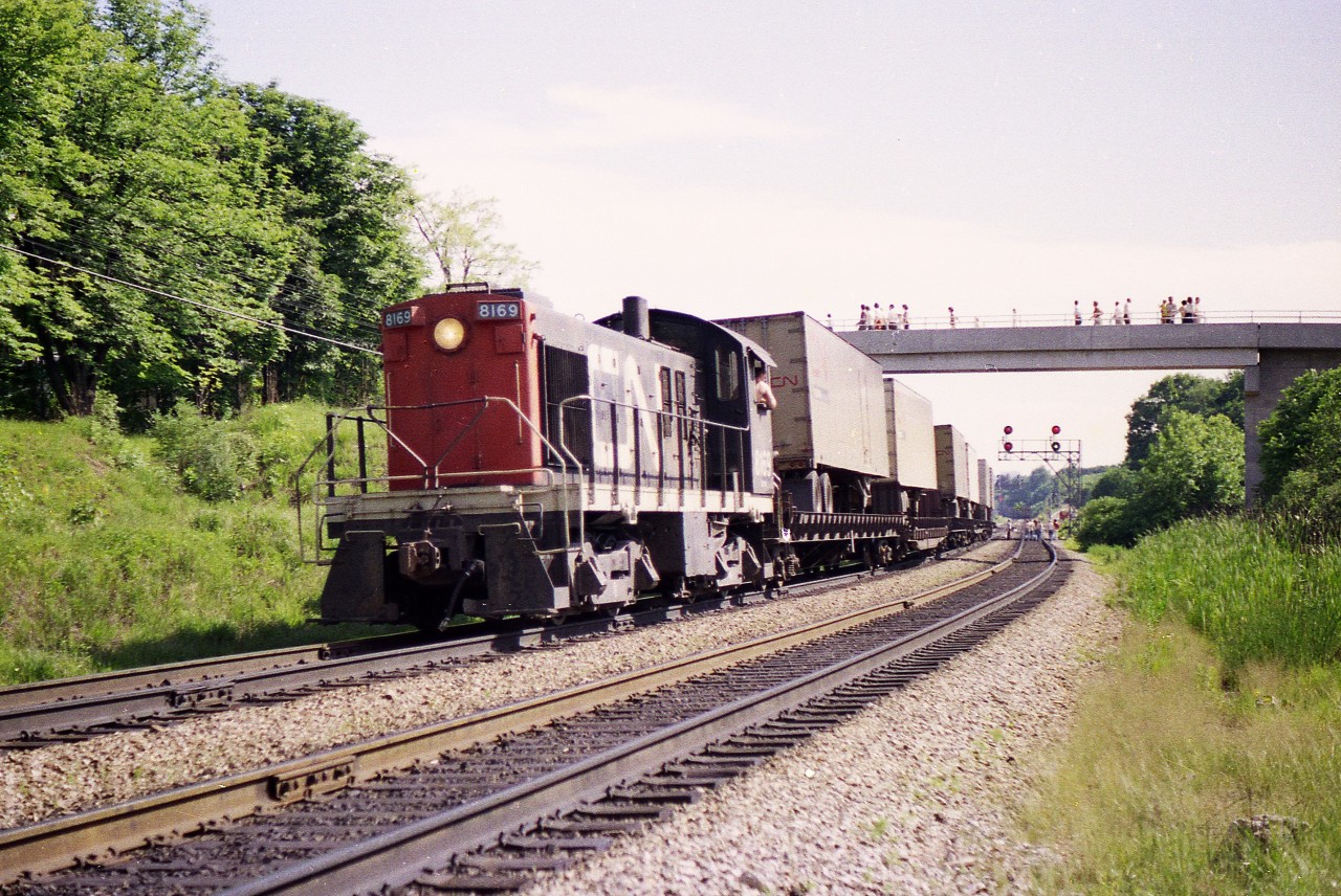 Many of us are frustrated by the strict control of property around the railroads. Fencing and No Trespassing signs, along with high quality cameras, are getting to be the norm. I thought posting this image might help clarify what has transpired over the years.
In this image, CN 8169, an MLW S-4, is in process of 'wyeing the pigs'. a daily (almost) procedure in which TOFCs are run around the wye in order to be in the correct position at the loading docks in Hamilton.
The engineer has just run the wye and he is now waiting for the signal in order to back the train into Hamilton.
Note there is a green signal on the right for a movement up the Dundas. Note the people on the tracks paying no attention whatsoever to potential danger on both tracks.
Railroads had to put up with this for years.   No more.