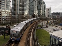 The Vancouver Sky train rounds a curve beneath towering multi-story apartments. A second train can be seen in the distance about to stop at a terminal. 