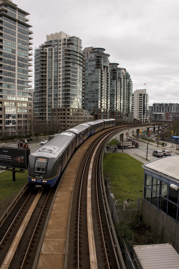 The Vancouver Sky train rounds a curve beneath towering multi-story apartments. A second train can be seen in the distance about to stop at a terminal.