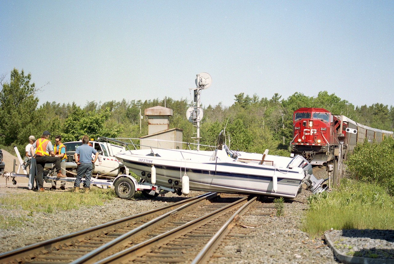 This was such an unusual incident.  I posted a photo more than 12 years ago of the train crews trying to push this boat off the track to no avail.  Figured this scene needed a revisit.
Luckily for all involved; a meet was to occur here, and the northbound train was just going into the siding and as a result the southbound pictured was moving rather slowly.
In this shot the boys are talking over what to do about this situation, (note the CP truck) and it turned out that all involved could not budge this boat, so the fork lift used by the boat storage place up on the hill came to the rescue.
I was just driving along and I heard the chatter on my scanner, which resulted in me showing up here. It appeared that, rolling over a very bumpy access road, the straps on the boat gave way and it slid off the trailer. I didn't ask questions.  My presence was not appreciated.
SB power in the photo  consists of a pair of SD90MAC, 9147 and 9153.