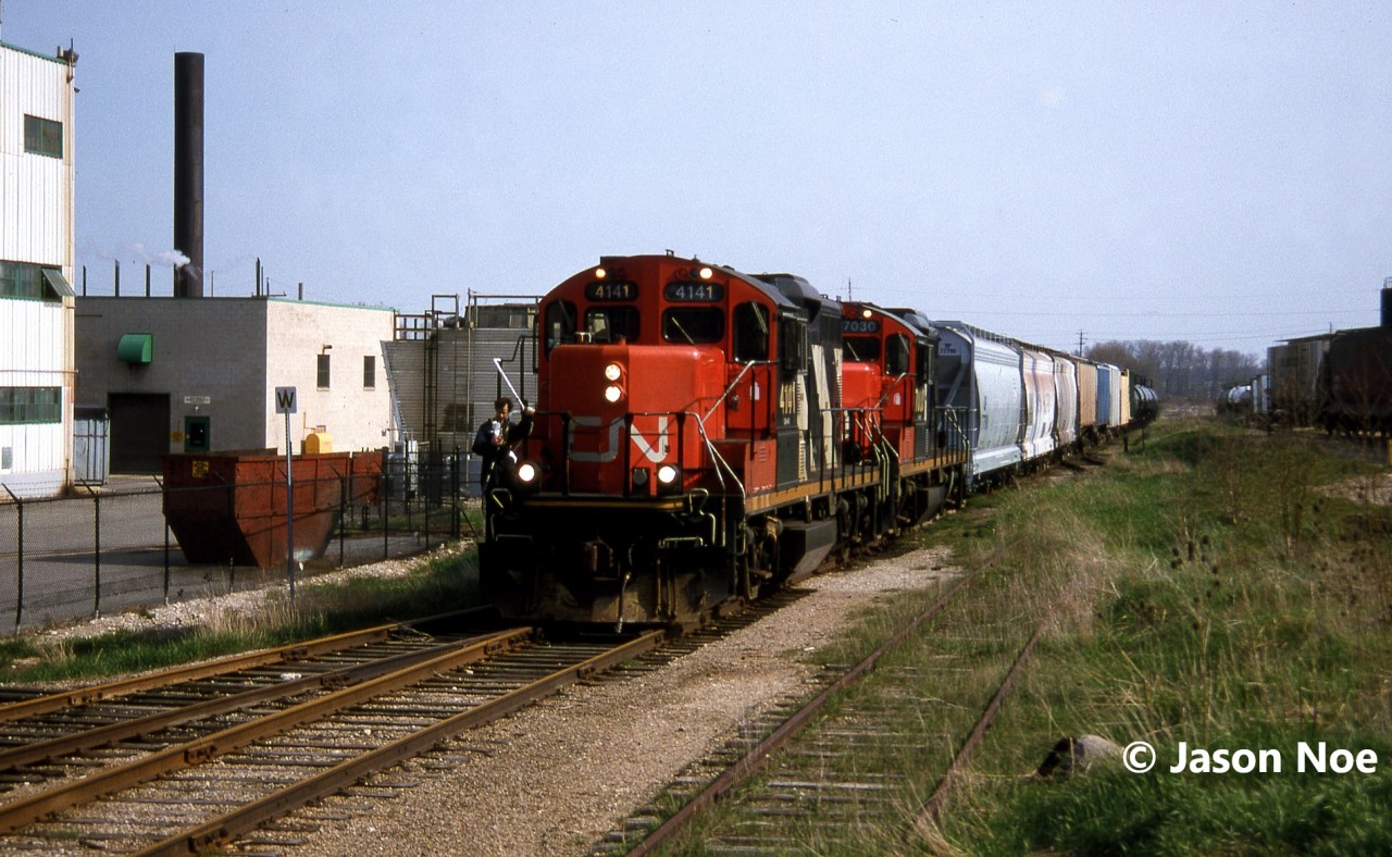 The CN 15:30 Kitchener Job is viewed arriving in Elmira, Ontario on the Waterloo Spur with a lengthy train and GP9RM’s 4141 and 7030. At the time, the job only operated with two units when it was necessary and on this day, both Nutrite and Sulco Chemicals in Elmira were receiving several cars. The springtime was always a busy time for Nutrite and their spur is seen to the right in the photo already full of hoppers previously set-off.