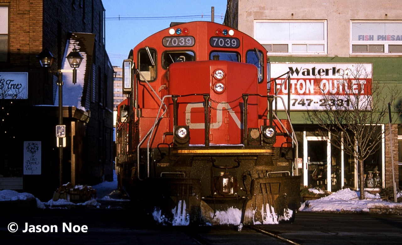 During a late winter evening, the CN 15:30 Kitchener Job is pictured crossing King Street in uptown Waterloo, Ontario with GP9RM’s 7040 and 7039. The units were returning to the Kitchener yard light power after setting-off tank cars in Elmira on the Waterloo Spur.