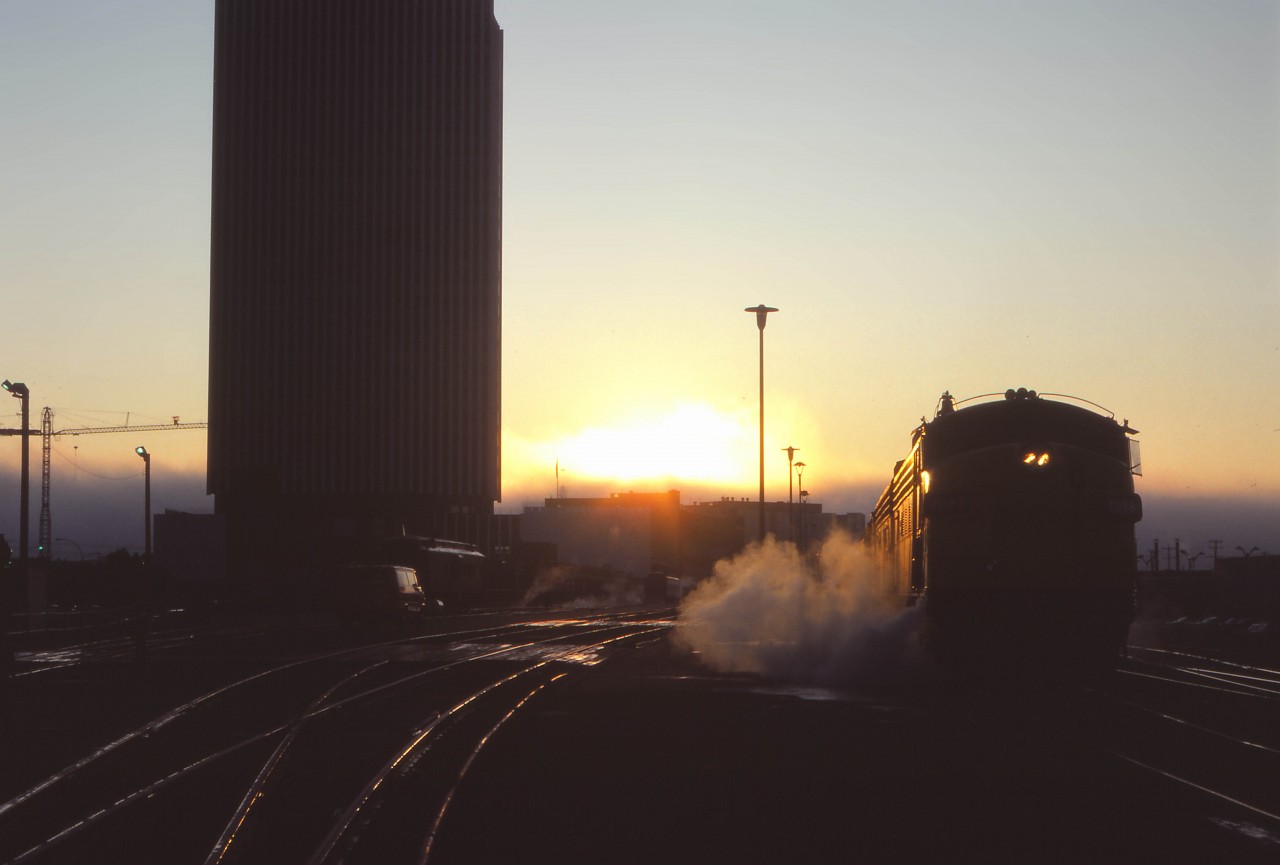 On what promises to be a nice September day, the sun rises on the westbound Super Continental being serviced in Edmonton. Alas, in a little over two months, “the Super” will no longer call here—replaced by day trains Winnipeg-Regina-Saskatoon, Saskatoon-Edmonton, and the “Rupert Rocket” extended to Edmonton. The passenger station was opened by Canadian National on 14 February 1966, and the CN Tower office building officially opened on 4 November 1966—at the time, the tallest office building in Western Canada.  Eighteen years after this photo was taken (on 3 September 1989), the restored Super Continental began the westbound pull-in/back-out move as CN phased out operations in downtown Edmonton. VIA would finally move to its current station near the Yellowhead Trail and Walker Yard on May 29, 1998. CN moved its office personnel out of the Tower a decade later.
