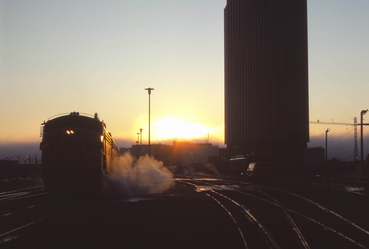 On what promises to be a nice September day, the sun rises on the westbound Super Continental  being serviced in Edmonton. Alas, in a little over two months, “the Super” will no longer call here—replaced by day trains between Winnipeg-Regina-Saskatoon, Saskatoon and Edmonton, as well as the “Rupert Rocket” extended to Edmonton. The passenger station was opened by Canadian National on 14 February 1966, and the CN Tower office building officially opened on 4 November 1966—at the time, the tallest office building in Western Canada.  Eighteen years after this photo was taken (on 3 September 1989), a restored Super Continental began the westbound pull-in/back-out move as CN phased out operations in downtown Edmonton. VIA would finally move to its current station near the Yellowhead Trail and Walker Yard on May 29, 1998. CN moved its office personnel out of the Tower a decade later. (Also note the heavyweight NAR passenger car next to the building.)