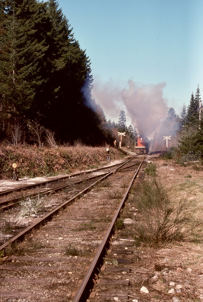 On Vancouver Island at Deerholme, CN 991 was left idling overnight when it was cold, causing excessive build-up in the exhaust system.  To minimize smoke exposure to the caboose crew on the next day’s work, at first move was a short run loading against full engine brakes, as seen here at the west end of the yard on Wednesday 1976–03-31.