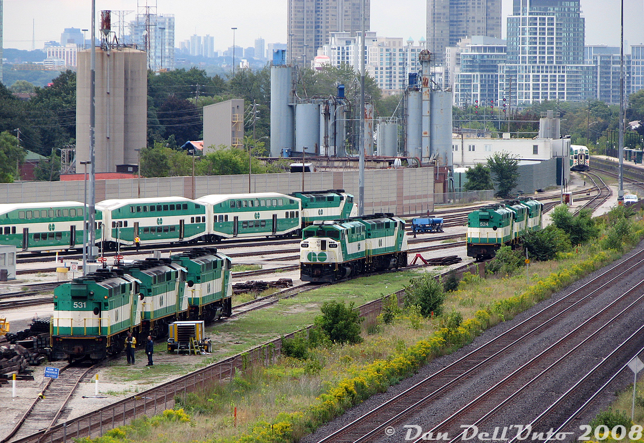 2008 on GO Transit was the beginning of the end of the F59PH fleet: as new MP40PH-3C units from MPI started to come online, old F59's started to populate the deadlines around Willowbrook Yard. Some were just stored out of service, while others had pushed their last bilevel. Units from the first order (520-535, built 1988) were the first to go: stored here are 531, 541 and 556 elephant style, the pair 532 and 520 back-to-back at the bumper, and 524, 533 and 521 shoved out of the way at the far end. It was very rare to see a unit facing west, but GO turned one or two units to serve as shop switchers around Willowbrook in their later years (a back-to-back pair even made it out in revenue service). Two employees work on the closest units under blue flag protection, perhaps assessing 533's condition for potential sale. 541 and 556 would return back to work for a year or two, but the rest sold between March-May 2009 as the deadlines grew more. Unit 563, still active, is pictured in the background getting ready to take out a PM rush hour train.

Other neat things in this photo are the low building behind the sound barrier wall belonging to Canadian Foundry Supplies (now demolished, they had their own siding that received hoppers into at least 2007). In the distance on the right is GO's Mimico station, and all the condos in the distance along the Lake Shore repopulated the old motel strip. If you eye-spy a bit, you can pick out the water tower for Mr. Christie's Lakeshore Bakery (still active, later demolished), and the blue load-test unit built from an EMD dynamic brake hatch.