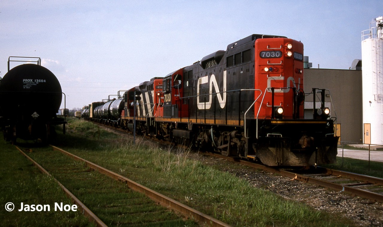 The CN 15:30 Kitchener Job is viewed switching in Elmira, Ontario on the Waterloo Spur with a lengthy train and GP9RM’s 7030 and 4141. At the time, the job only operated with two units when it was necessary and on this day, both Nutrite and Sulco Chemicals in Elmira were receiving several cars. The springtime was always a busy period for Nutrite receiving hoppers. Also, the crew had a loaded lumber car that they would set-off at Fairway Lumber in St. Jacobs on their return trip south to Kitchener.