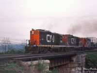 The engineer of this eastbound CN freight throttles up his three GP9 units as they motor through Bayview Junction in the evening. CN 4516, 4575, and 4588 lead with some piggyback traffic up front (with a pair of Imperial Roadways "Puptainer Service" trailers riding at the head end).<br><br>The CN-style spark arrestors on the roof gave CN's GP9 units (and other GMD units with them) a distinct exhaust note, sounding like dull thumping on the side of a steel drum. This carried over to the rebuilt units that retained them. <br><br><i>Bill McArthur photo, Dan Dell'Unto collection slide.</i>