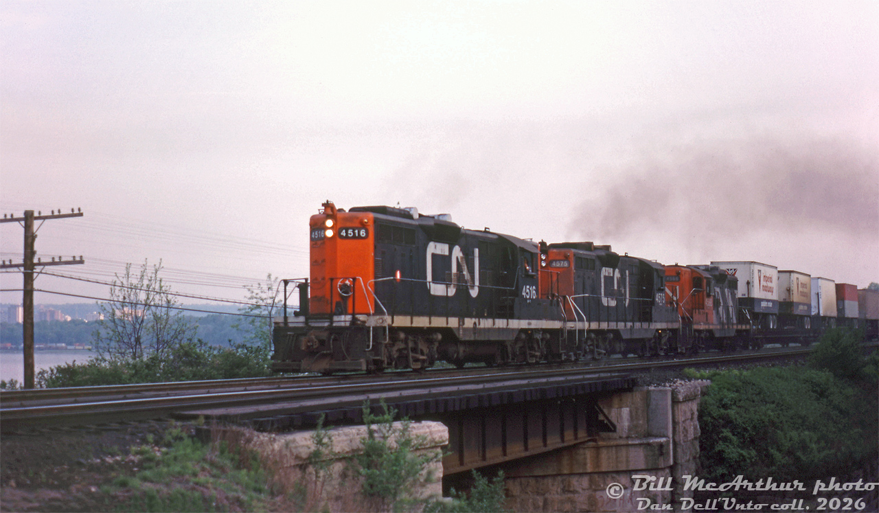 The engineer of this eastbound CN freight throttles up his three GP9 units as they motor through Bayview Junction in the evening. CN 4516, 4575, and 4588 lead with some piggyback traffic up front (with a pair of Imperial Roadways "Puptainer Service" trailers riding at the head end).

The CN-style spark arrestors on the roof gave CN's GP9 units (and other GMD units with them) a distinct exhaust note, sounding like dull thumping on the side of a steel drum. This carried over to the rebuilt units that retained them. 

Bill McArthur photo, Dan Dell'Unto collection slide.