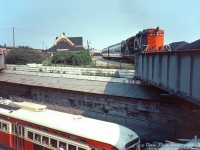 A rather unconventional angle of Parkdale, with a few photographic faux-paux, but full of all sorts of rich content: CN GP9 4518 (a freight unit) pulling today's "Barrie Bullet" commuter train for the <a href=http://www.railpictures.ca/?attachment_id=44939><b>Newmarket Sub</b></a>, is seen making its station stop at CN's Parkdale Station (replaced with a <a href=https://www.railpictures.ca/?attachment_id=58649><b>CN insta-shack</b></a> a few years later). To the left is CN's old <a href=http://www.railpictures.ca/?attachment_id=43465><b>Parkdale freight shed</b></a>. To the upper right is the roof of CP's own Parkdale Station building (closed a few years earlier in 1968) and <a href=http://www.railpictures.ca/?attachment_id=53581><b>yards</b></a> visible in the distance. The main CN Weston Sub crossed the steel girder bridge on the right, the one on the left had a service track running north to <a href=http://www.railpictures.ca/?attachment_id=45492><b>CN's small yard and facilities</b></a> nearby.<br><br>Below, traffic passes by on the busy <a href=http://www.railpictures.ca/?attachment_id=51378><b>Queen Street West</b></a>, including TTC PCC streetcar 4485 on the Queen route. On the left site along the brick retaining wall under the awning, one can see the opening and stairway that led up from Queen Street to CN's Parkdale Station above (there was a <a href=http://www.railpictures.ca/?attachment_id=31661><b>similar stairway</b></a> to access CP's Parkdale Station off Dufferin Street).<br><br>This neat photo was taken by the same unknown Toronto-area photographer who took <a href=https://www.railpictures.ca/?attachment_id=36087><b>this shot of GO 9807</b></a> from a slightly different angle, on the same day, in the same spot.<br><br><i>Original photographer unknown, Dan Dell'Unto collection slide.</i>