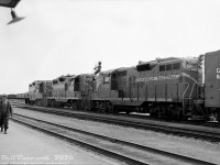 Three of CN's plentiful GP9 units, 4463, 4403 and 4600, sit in front of the station at Danforth in east-end Toronto on an eastbound freight as a member of the crew walks the platform, appearing to be eyeing the station. It's April 1961, and the last steam engines on the system had just been retired the previous year in favour of hoards of first-generation diesels such as GMD's popular GP9 model. Notes by the unknown photographer of this negative indicate the train was of ample size at 103 cars long, which would likely have required a helper engine for the eastbound grade here during the steam era. Freight cars visible in the background are parked in the adjacent Danforth Yard, CN's main east-end Toronto freight yard that would hold importance until its gradual decline after the "Toronto Bypass" and Toronto (MacMillan) Yard opened to the north of the city. Today the yard has been redeveloped into housing, and this location is now home to Danforth GO Station.<br><br>For the modelers out there, CN's 44/45/4600-series GP9's were regular or "heavy" freight units, as opposed to the light-weight 41/42/4300-series units. 4403 is one of the earliest such GP9's, exhibiting the old Pyle reflector style or "ash can" barrel headlights, a Phase 1 carbody configuration and early bolted stanchions. The other two units are later Phase 2 GP9's, but feature a unique-to-CN carbody louvre arrangement that moved one of the 2x3 set onto the next door, in order to spell out the full "Canadian National" lettering clearly. These units all had smaller 1000 gallon (imperial) fuel tanks, versus the more common 1333 gallon (imp, or 1600 US gallons) found on other railways. Rooftop-mounted spark arrestors and a long hood mounted bell are typical CN fare. In a way it's fitting that similar-looking CN GP7 4803 is preserved at the Toronto Railway Museum, a good representation of all the early CN Geeps that once handled freight in the post-dieselization era.<br><br><i>Original photographer unknown, Dan Dell'Unto collection negative.</i>