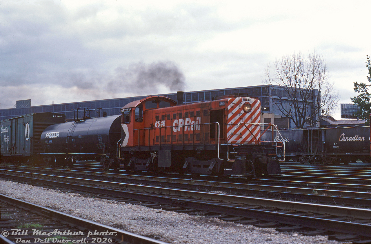 The engineer of CP Rail S3 6545 notches the little unit's throttle up, working around CP's Parkdale Yard in the Liberty Village area of Toronto. In the background behind the yard tracks is the John Inglis appliance factory, and other industrial buildings that once populated this area. 6545 was just one of many little steam era Alco/MLW S2/S3/S4 diesel switchers CP employed to handle industrial and switching duties in and around the Toronto area. In consist is extended-height CP newsprint boxcar 84981, increased in size to hold newsprint rolls stacked double high.

This area is now heavily gentrified, the last of the Inglis plant (and its iconic billboard sign) have been demolished, and the rail corridor through here is now a giant trench ducking under Strachan Avenue. The last of CP's little Alco/MLW switchers were retired in the mid-1980's with the closing of CP's John St. Roundhouse downtown.

Bill McArthur photo, Dan Dell'Unto collection slide.