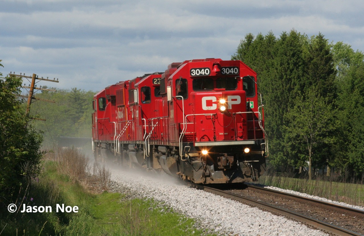 An extra CPKC Wolverton Job with GP38-2 3040 and GP20C-ECO's 2307 and 2259 is pictured heading westbound at Mile 65 on the Galt Subdivision, approaching Ayr, Ontario. The crew is returning to Wolverton after setting-off auto racks on the Waterloo Subdivision for the Toyota Plant in Cambridge, with a stop at Ayr ahead to switch FS Partners on the Ayr Pit Spur. June 1, 2025.