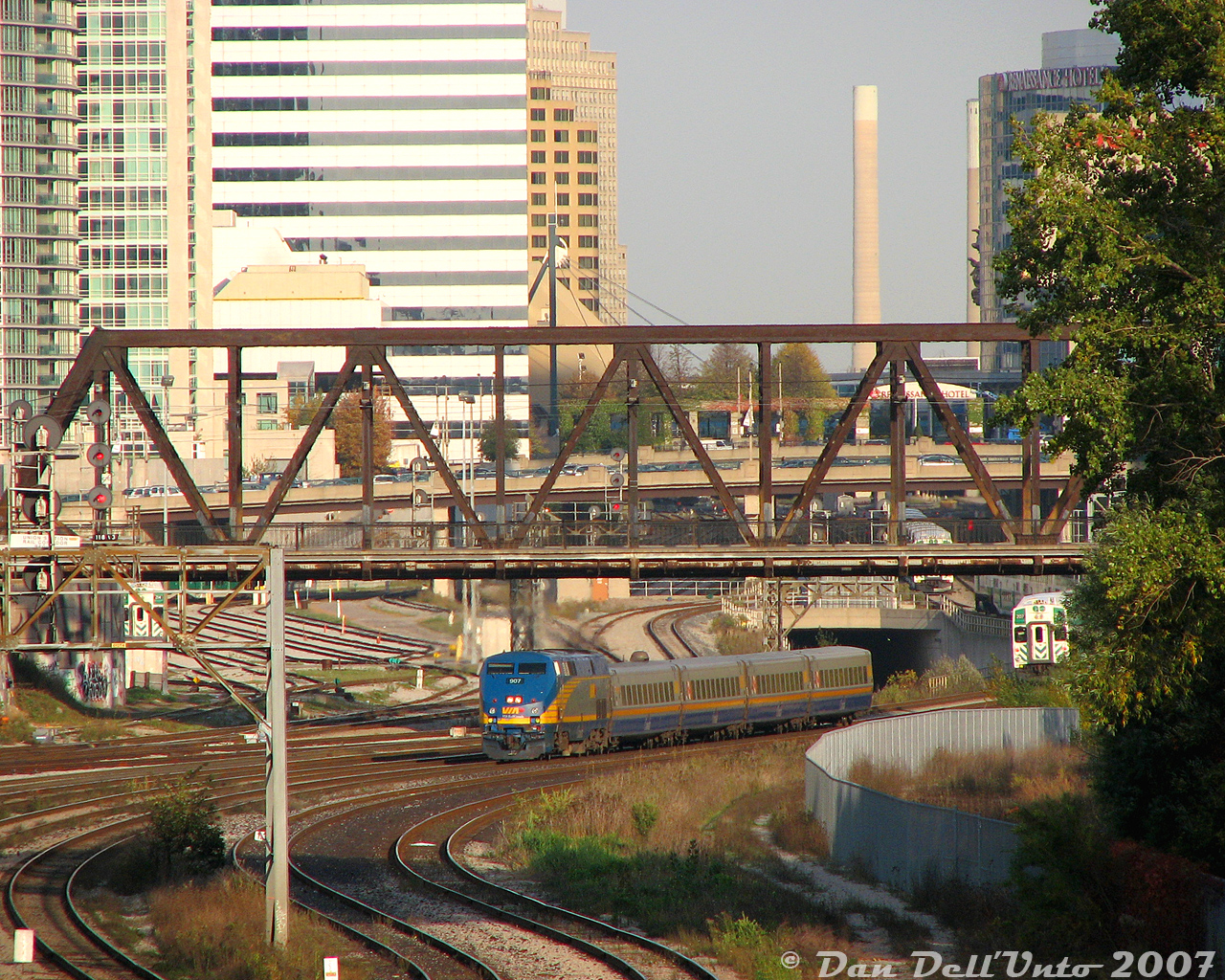 Not the conventional Bathurst Street bridge shot: this one is of the bridge itself, looking east from Strachan Avenue in nearby Liberty Village.

VIA P42DC 907 backs its 4-car LRC consist from the TMC in Mimico to Union Station in downtown Toronto, ducking underneath Bathurst Street bridge after having just entered the Union Station Rail Corridor at the signalbridge sign marker to the left. GO trains are hiding in the wings: a few consists are lined up on the right, slowly making their way to Union Station to enter PM rush-hour service. At least one lingers in Bathurst North Yard beyond the bridge to the left, almost all cleared out of consists.

Once #87 arrives at Union and finishes boarding, it will make its timely departure from Toronto at 5:40pm (5 minutes ahead of Georgetown-line train #211 running behind it), bound for such exotic places as Brampton, Guelph, London, and ultimately Sarnia.