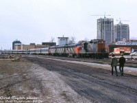 Back in the era of long passenger trains with the power to match, CN 4103, one of the passenger GP9's with high-speed gearing, leads an A-B-A set of FPA4 6770, a 6600-series F9B and 6500-series FP9 on an eastbound passenger train, at least 12 cars long, leaving London Station (the black office tower of which is visible in the distance). The units have just crossing Colbourne Street, with two pedestrians leasurely strolling into view alongside the track.<br><br><i>Gord Taylor photo, Dan Dell'Unto collection slide.</i>