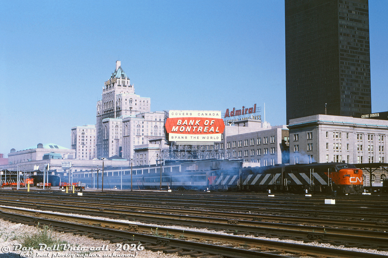 Pulling out of Union Station in downtown Toronto, CN's eastbound "Lake Shore Limited" departs from Track 2 with an A-B-A set of cowl units providing the power today. CN FPA4 6774, F9B 6620, and FPA4 678? start off the journey with some white smoke, after idling awaiting their departure time. CP Rail's (CP Hotels) Royal York Hotel can be seen prominently in the background across from the main Union Station building, with the Dominion Public Building on the right, almost all matching in colour. Behind the train are the old CP Express sheds (eventually demolished for the 2000's GO Transit bus terminal). One of the striking new black TD towers can be seen, the Toronto skyline not yet populated with a wall of office towers and high rises. That "Spans the World" Bank of Montreal billboard on top of the CP Express building is reminiscent of a similar old CP slogan from the 1940's-50's.

Original photographer unknown, Dan Dell'Unto collection slide.