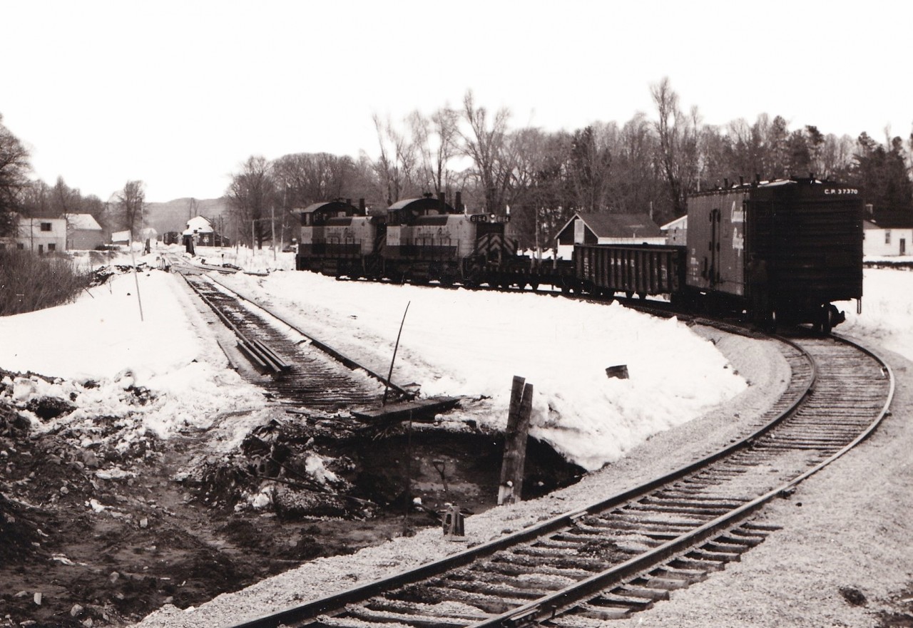 With a brakeman riding the rear, a pair of SW1200RS units take a few cars around the newly completed diversion track between the CNR Coldwater spur and the remnant of the CPR Port McNicoll subdivision.

This connecting track permitted CP to reach the CNR Midland sub at Coldwater when the large Hoggs Bay trestle was abandoned in 1971. A similar connecting track was built between McMillan on the Midland sub and the yard at Port. Prior to this CP had an interlocked  diamond crossing of the CN spur. CP used trackage rights on CN to reach McMillan until 1991 when the Port McNicol sub was abandoned. The diamond remained in place for several years until the unused rails were lifted between this point and the trestle.

The connecting track originally dipped south of the existing track before connecting to the existing track near the bridge over the Coldwater river. The connecting track was later reconfigured to a more conventional turn in the 1980's due to difficulties with long trains. Six axle units were prohibited on this later track arrangement. Our family rode a UCRS excursion to Midland that went through this trackage in 1975. It was an interesting sight watching the 80 foot long coaches negotiate the shoofly.

Visible in the background is the CPR Coldwater station with some work service cars in the siding. Between the bridge and flanger sign is visible one of the ancient crossing signs with a danger light on top of the post. This signal is now displayed at the Coldwater Museum.

The County of Simcoe mapping website (https://opengis.simcoe.ca/public/) has aerial views of this area. The 1978 and 1983 views show the two different layouts.

Unknown photographer from my collection.