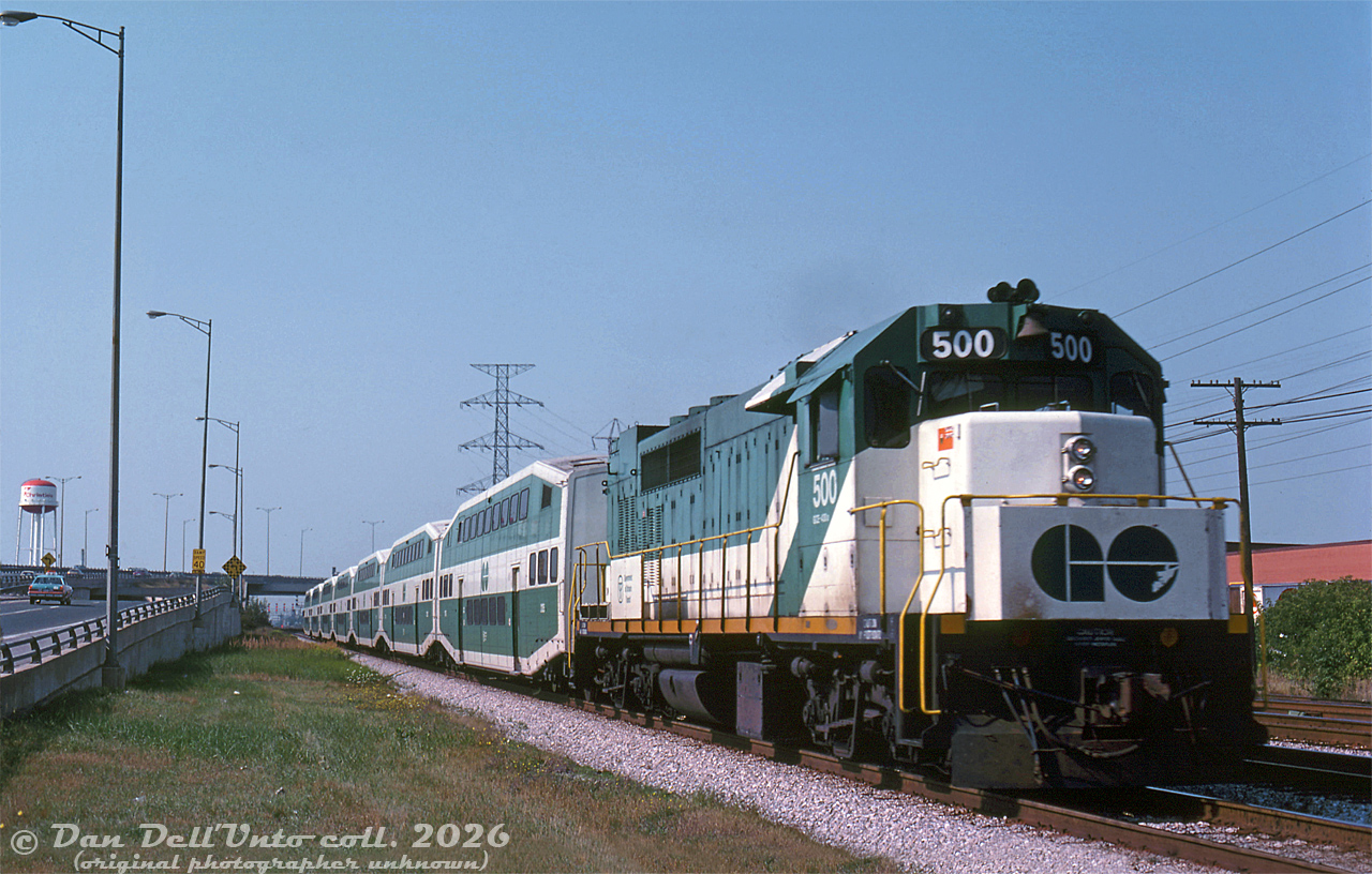 Heading eastbound into the city after dipping under the Queen Elizabeth Way overpass, GO Transit GP40TC 500 speeds through the Swansea area of Toronto with a 7 bilevel Lakeshore West train bound for Union Station. One can see the water tower for the Mr. Christie Lakeshore Bakery in the background, and part of the Oshawa Group warehouse (next to the Ontario Food Terminal) on the right. The signals for the Mimico East interlocking are visible in the distance.

Original photographer unknown, Dan Dell'Unto collection slide.