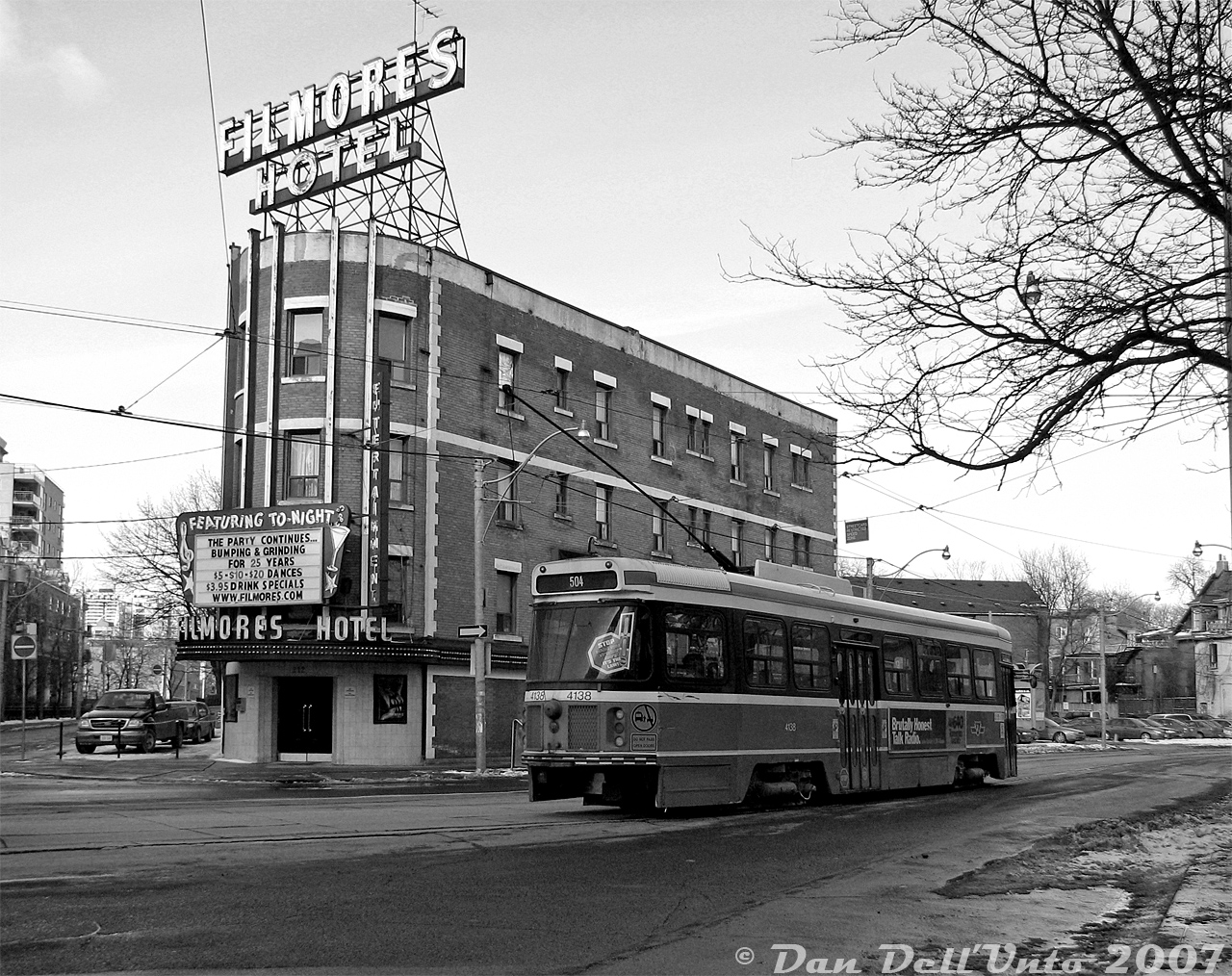 On a somewhat overcast winter's afternoon, TTC CLRV 4138 on the 505 Dundas rumbles by Filmore's Hotel, the infamous Toronto strip club with a hotel above it (they really do operate a hotel there, although how much sleeping really goes on inside is questionable). Features advertised to welcome passerbys such as us include "Bumping and Grinding", a selection of affordable dance rates with the lovely ladies inside, and drink specials for those so inclined to wet their whistle (note: no shenanigans or debauchery was partaken on this day).

If I was a Ryerson Image Arts Student, I would call this my B&W film period, and instead be wandering around West Queen West eating an avocado toast from Starbucks while shooting manhole covers, construction pylons, and various types of roadkill as still art, rather than be wandering around the seedy underbelly of downtown shooting streetcars. Unlike the Imperial Pub that was in the "safe" area of Dundas, once you wandered further east of the Ryerson campus and passed the Howard Johnson's at Jarvis Street, things started getting sketchy. Filmores was like a big sign, in more ways than one, warning you you weren't in that tourist-friendly Dundas Square/Eatons Centre area anymore. This area around Dundas, Jarvis and Sherbourne had a number of problems dating from the 1990's including crack cocaine, prostitutes, johns, transcients, drug dealers, addicts, homeless, flophouses and such. There's an informative NFB documentary called "East Side Showdown" (1999) that documents some of the shady goings-on in this area. It didn't seem to bad during the day during this admittedly brief foray, and in recent years, gentrification and new developments have taken most of the edge off.

Research shows the hotel building itself dates from the late 1800's, but the "gentlemen's club" opened sometime in the early 1980's. The property, located at the jog in Dundas at St. George, was sold to developers for potential redevelopment during the pandemic, but the strip club portion continued to operate until it closed sometime in 2026 (the hotel portion appears to still be open for now). Being one of those "unique" places that gives certain neighbourhoods in Toronto their character, it's likely in the near future to be replaced by a bland condo development with a few chain retail stores at ground level (and it's likely the ladies workin' the new Shoppers Drug Mart can't compare).