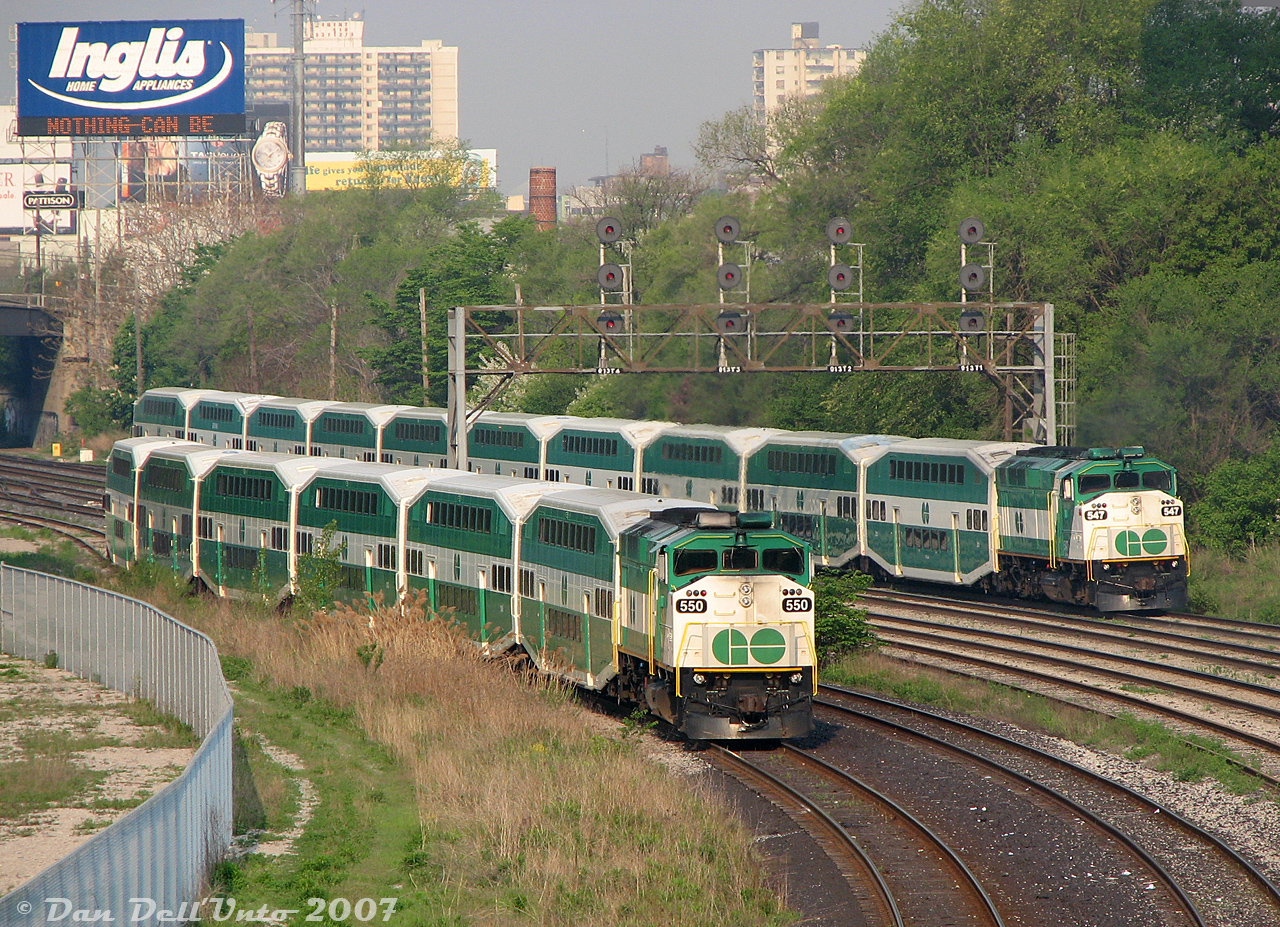Little Train, Big Train: a pair of GO Transit F59PH units handle two morning rush hour trains leaving the Union Station Rail Corridor onto the CN Oakville Sub near Strachan Avenue: GO 550 off Bradford (Barrie) line train #802 pauses with its short equipment move waiting for the light off the bypass/"High Line" tracks to proceed west to Willowbrook Yard, passed by in-service 547 buzzing by at speed on the tail-end of full-sized Lakeshore West train #477.The ever-present Inglis sign offers its morning insights of "Nothing can be changed until you start(?) to find the issue", that smokestack belongs to one of the old restored buildings in Liberty Village, and I believe those apartment buildings are off Dufferin Street.At the time, GO's smallest consists were "L6" trains (locomotive and 6 bilevels), the smallest possible to keep the required 2500-series "5A" accessible car as the 5th car in order to line up correctly with the accessibility platforms. Some Georgetown, Bradford and other lines used them on less-patronized trips, while the standard "L10" consist (locomotive and 10 bilevels) was the maximum-sized train GO would run to do the heavy lifting on busier trips. At the time GO was all-F59 all the time, and their HEP abilities limited trains to a maximum of 10 cars (most station platforms were also only that long).