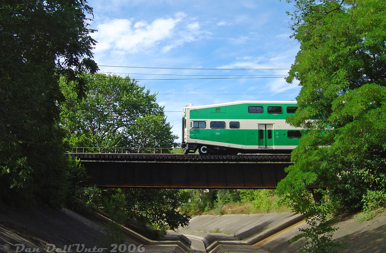 GO Transit cab car 210 punches out of the foliage above the Etobicoke Creek, heading west on train #281 (Union-Bramalea with F59 563 on the rear) on the final push to downtown Brampton over the single track section of the Halton Sub between Peel and Brampton East. We had just made it over here from the Dixie Cup Spur to shoot CN #559 (finished from switching Georgia-Pacific) backing past and slipping into the clear at Peel about 10 minutes before. Shooting anything off the pedestrian bridge here, you had to keep your ears peeled and know when something was coming in order to time the shot correctly, since there was no visibility down either side of the line. The regular GO trains with a schedule committed to memory made things a bit easier.

For some very not smart reason lost to time, the old town of Brampton (downtown) was built OVER the Etobicoke Creek, with the original alignment following Main Street winding south underneath some of the buildings downtown. This caused many seasonal flooding issues, including very significant flooding of the entire downtown area in March 1948 (see olde-timey documentary here, 10:32 shows this exact spot). After this, Brampton had enough of decades of mother nature casually trying flood it out of existence. A new concrete diversion channel was built to the east to divert the creek alignment out of and around the downtown area, running from Vodden Street to here, just south of a new CN bridge. At the time of this photo, the bridge was part of the single-track "gauntlet" between http://www.railpictures.ca/?attachment_id=9714Brampton East and Peel, but CN would soon add another span here in 2006-2007 on the north side in order to double-track the line all the way through.