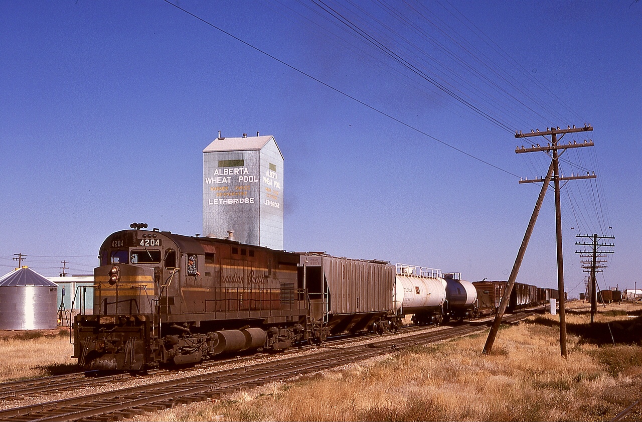 Timetable train No. 83 was a wayfreight between Medicine Hat and Lethbridge, shown here arriving near the east end of Lethbridge at 1430 MDT on Thursday 1974-10-03 when the line still ran through downtown.  I have seen many filthy locomotives, and C-424 CP 4204 certainly deserves a prize — imagine the river of muddy water resulting when it finally was washed!